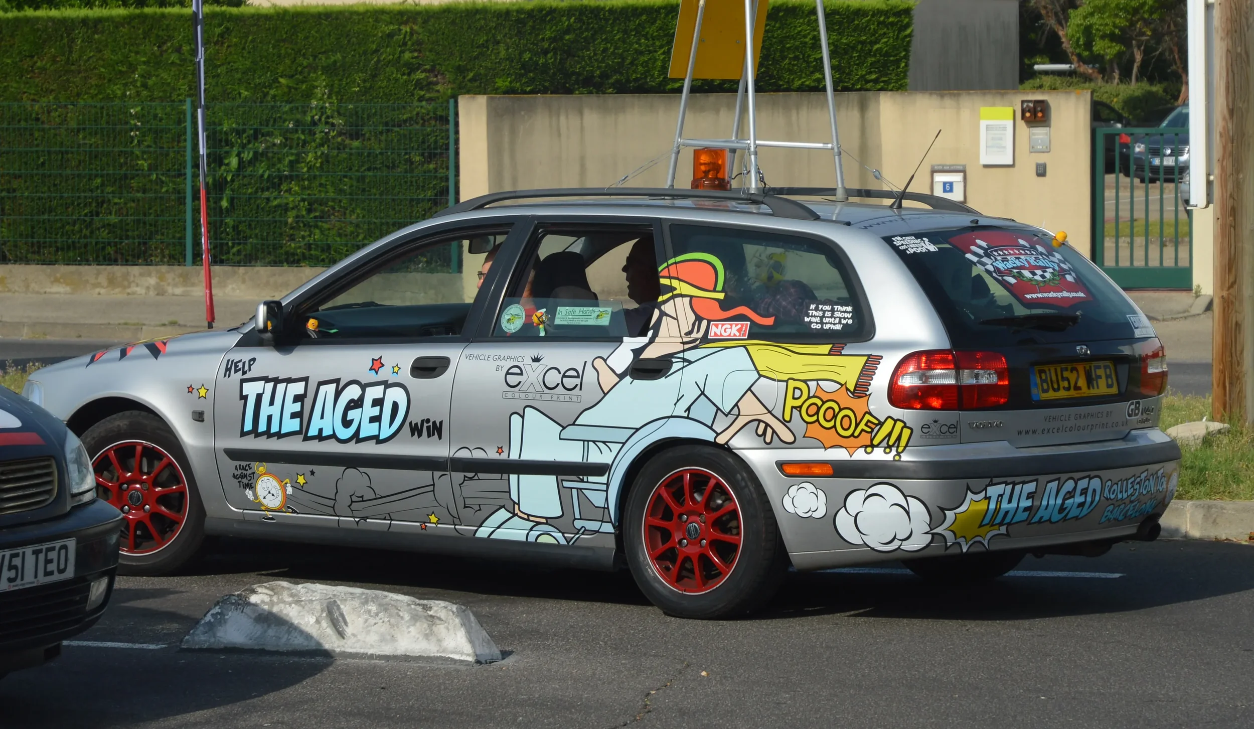 A silver car decorated with comic-style graphics and text, featuring a cartoon character of an older person with sunglasses and a cap, along with phrases like "Help The Aged Win," "Pcoof!!," and "Rolling Stone Backstage." The car has red rims and is 