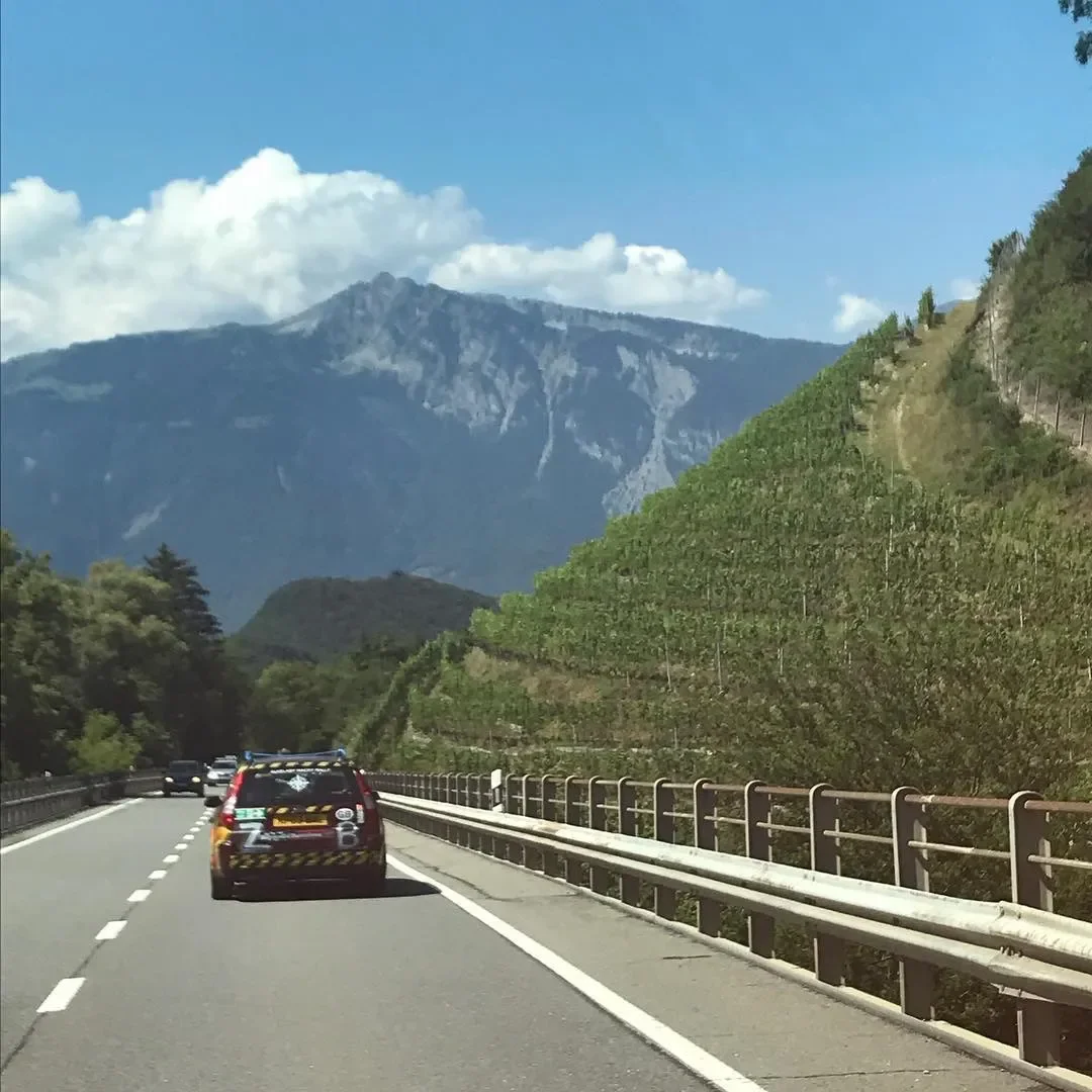 A scenic view of a mountain road with a black SUV driving along a winding highway, surrounded by green hillsides and a mountain in the background under a partly cloudy sky.
