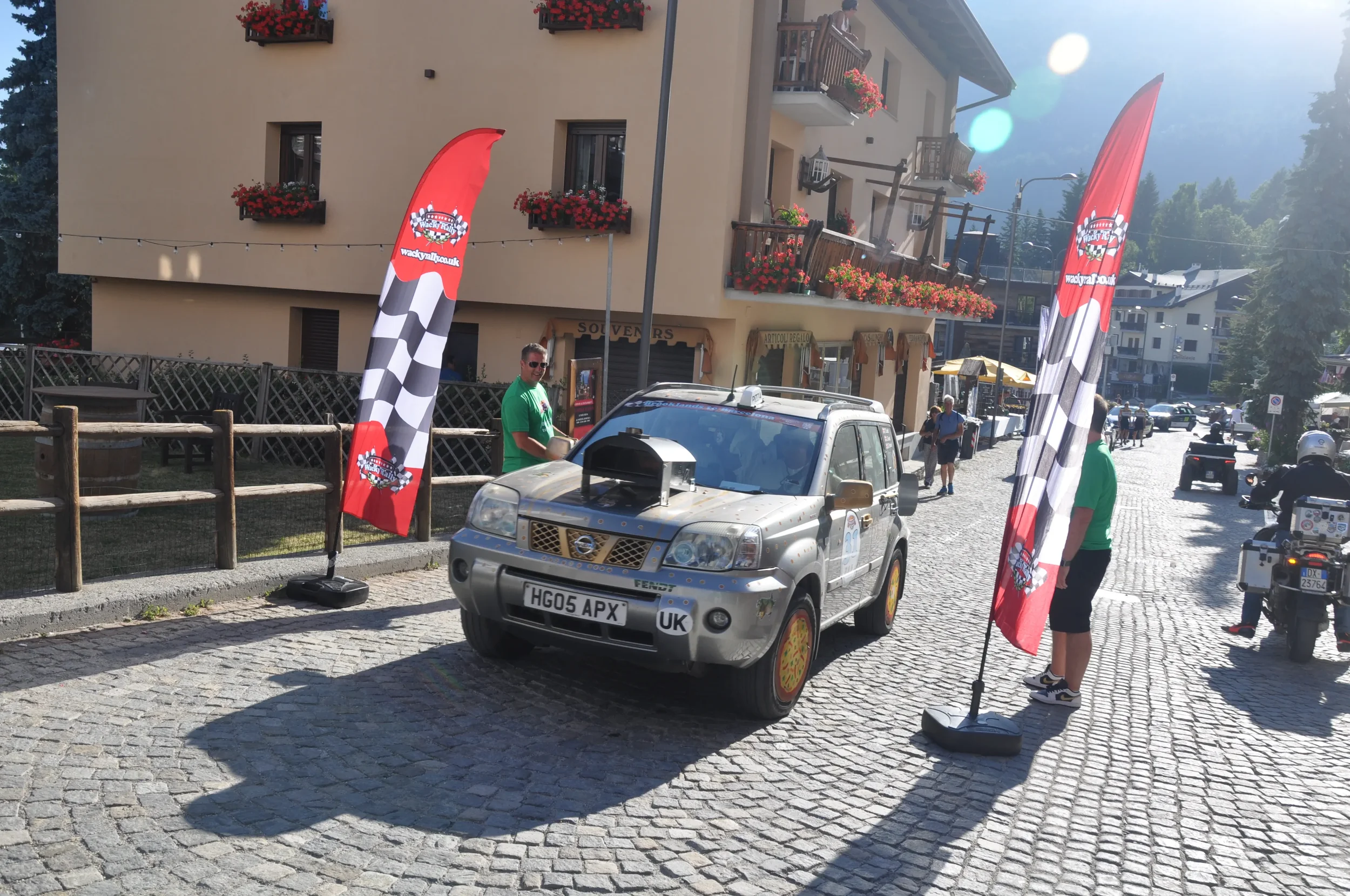 A small rally car with a pizza slice sticker on the wheel hub parked on a cobblestone street, flanked by two checkered flags with red, black, and white logos and the website 'wackyrici.uk'. Two people wearing green shirts are standing nearby, one on 