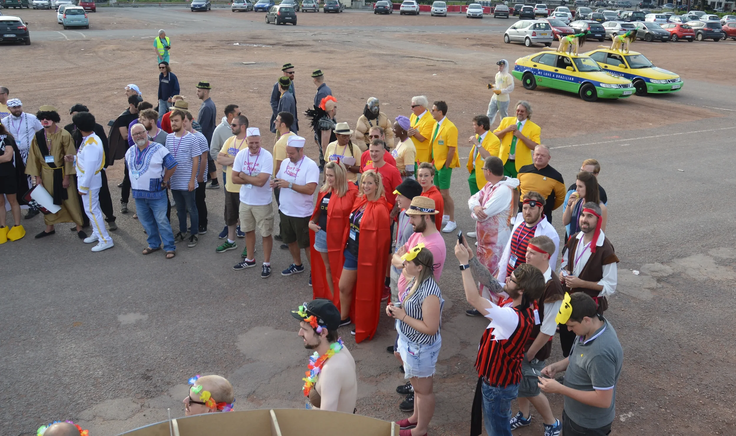Group of people dressed in colorful costumes and accessories standing in an outdoor parking lot, some taking photos. In the background, there are cars, yellow and green taxis, and people in casual attire.