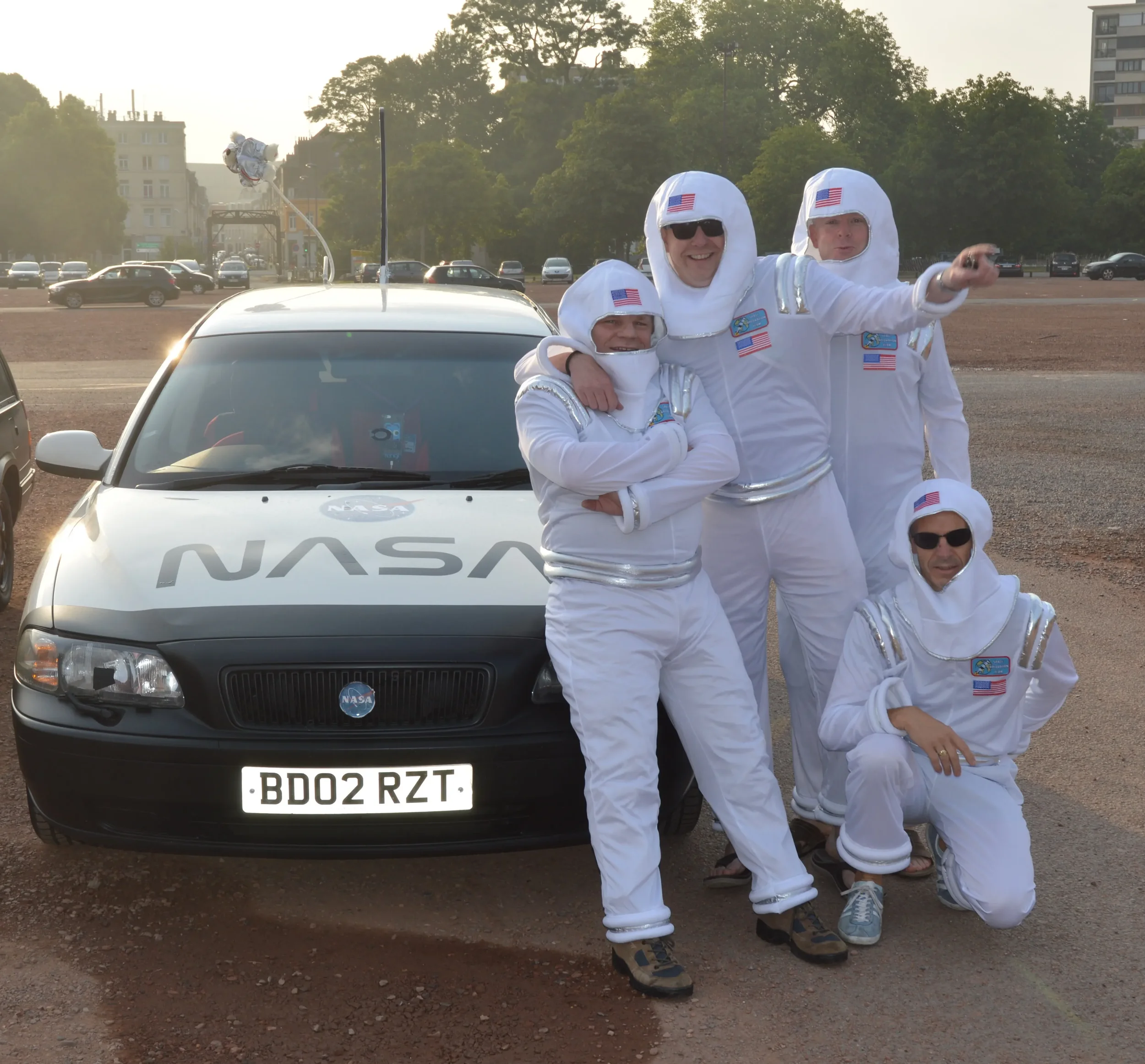 Six people dressed in astronaut costumes standing beside a NASA-branded car in an open parking lot, with a cityscape and trees in the background.
