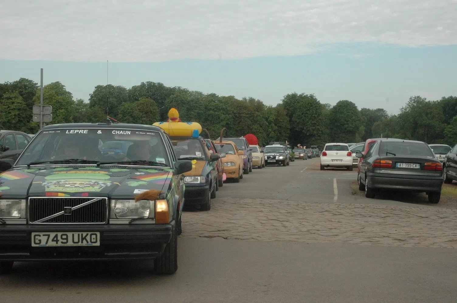 A parking lot filled with various cars, including a decorated black Volvo with a Louisiana laser tag truck-themed design, an inflatable yellow duck on top of a car, and a line of cars extending into the distance, surrounded by trees under a cloudy sk