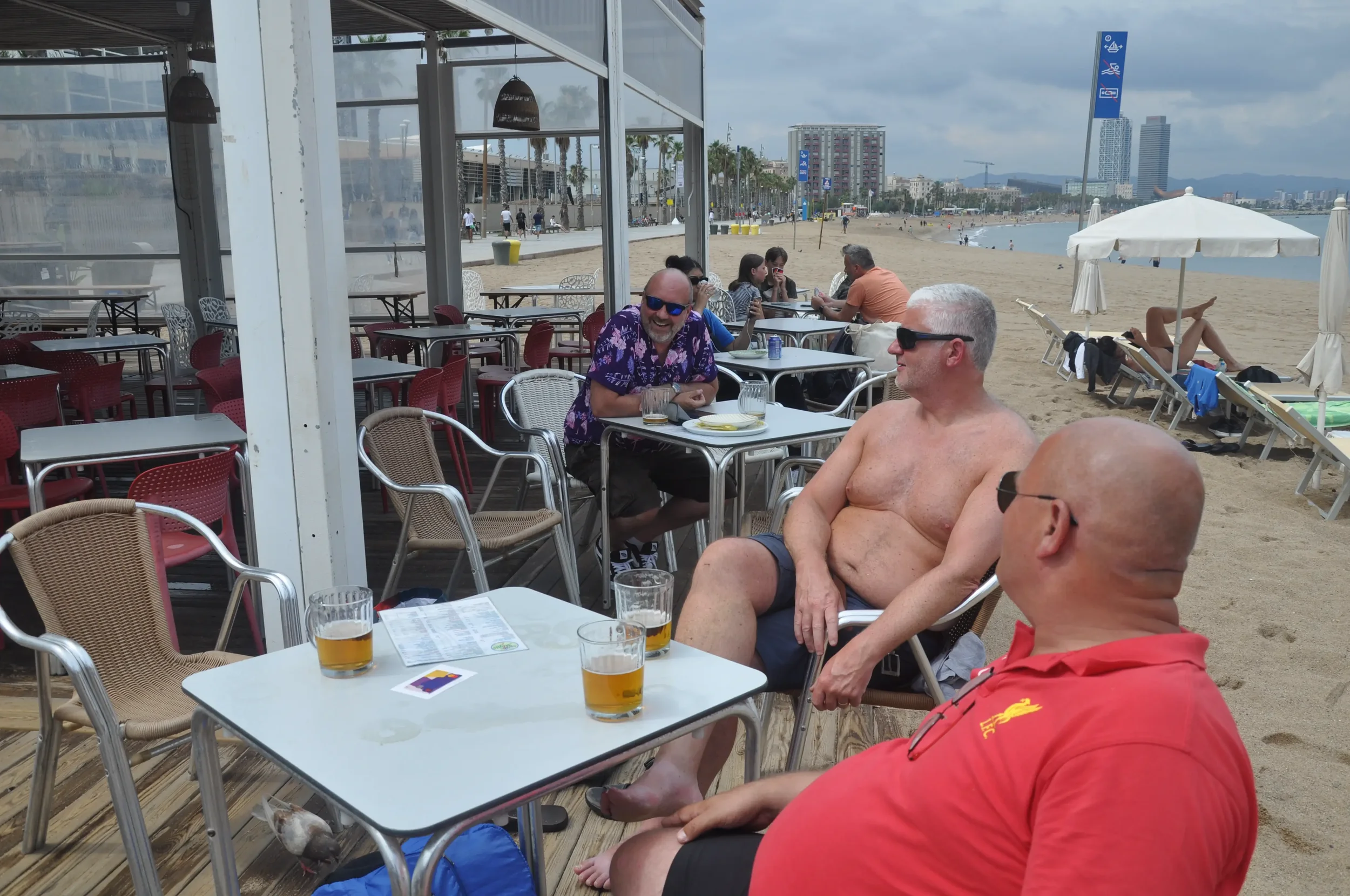 Three men sitting at a table on the beach, one shirtless and two wearing sunglasses, with drinks in front of them. There are empty tables and chairs, beach umbrellas, and people in the background along the shoreline.
