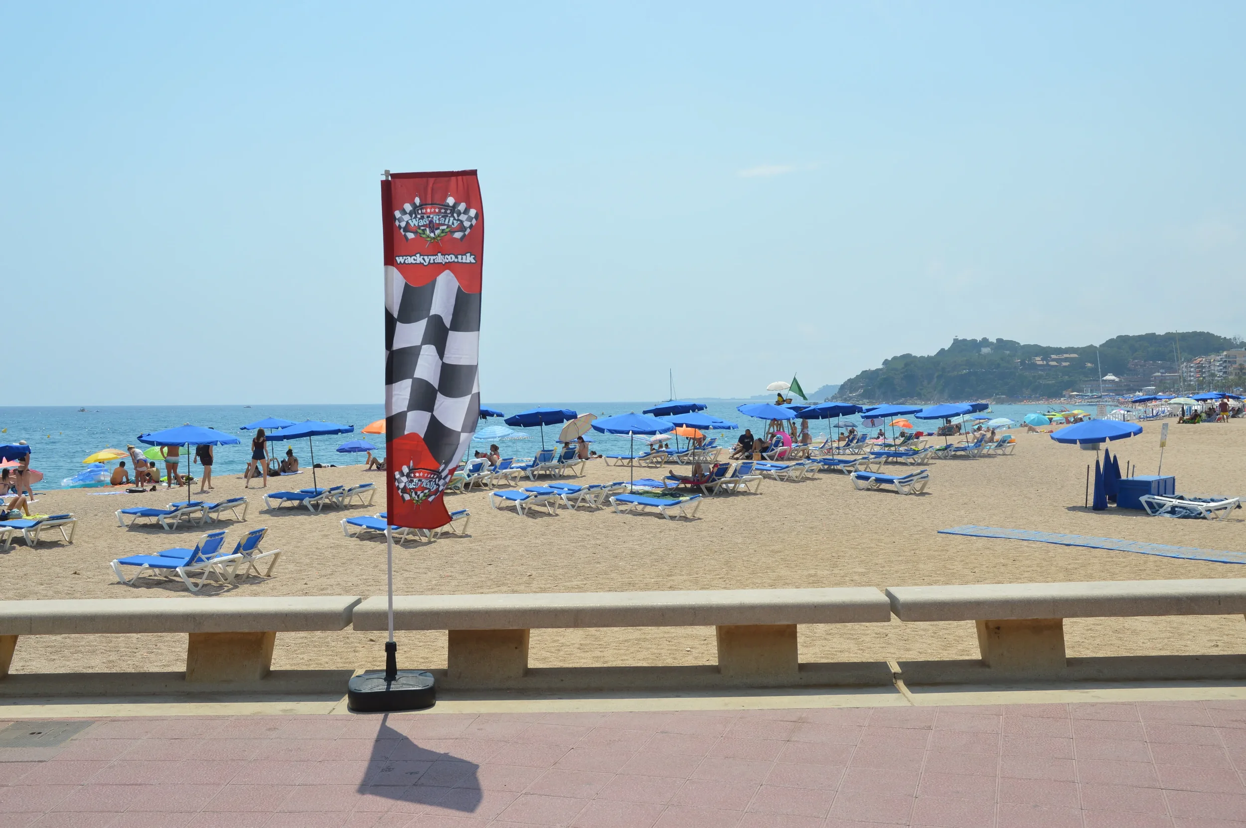 Beach scene with blue umbrellas and lounge chairs on sand, people near shoreline, and a red flag with checkered pattern and racing logo. Hills in background and a cityscape to the right.