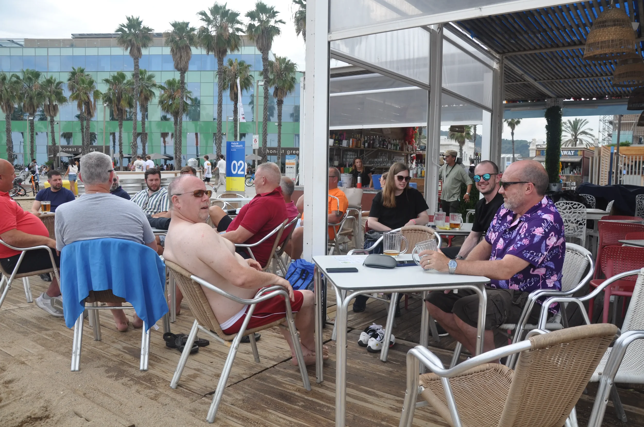 People sitting at outdoor tables near the beach, with palm trees and a colorful building in the background, enjoying drinks and conversation.