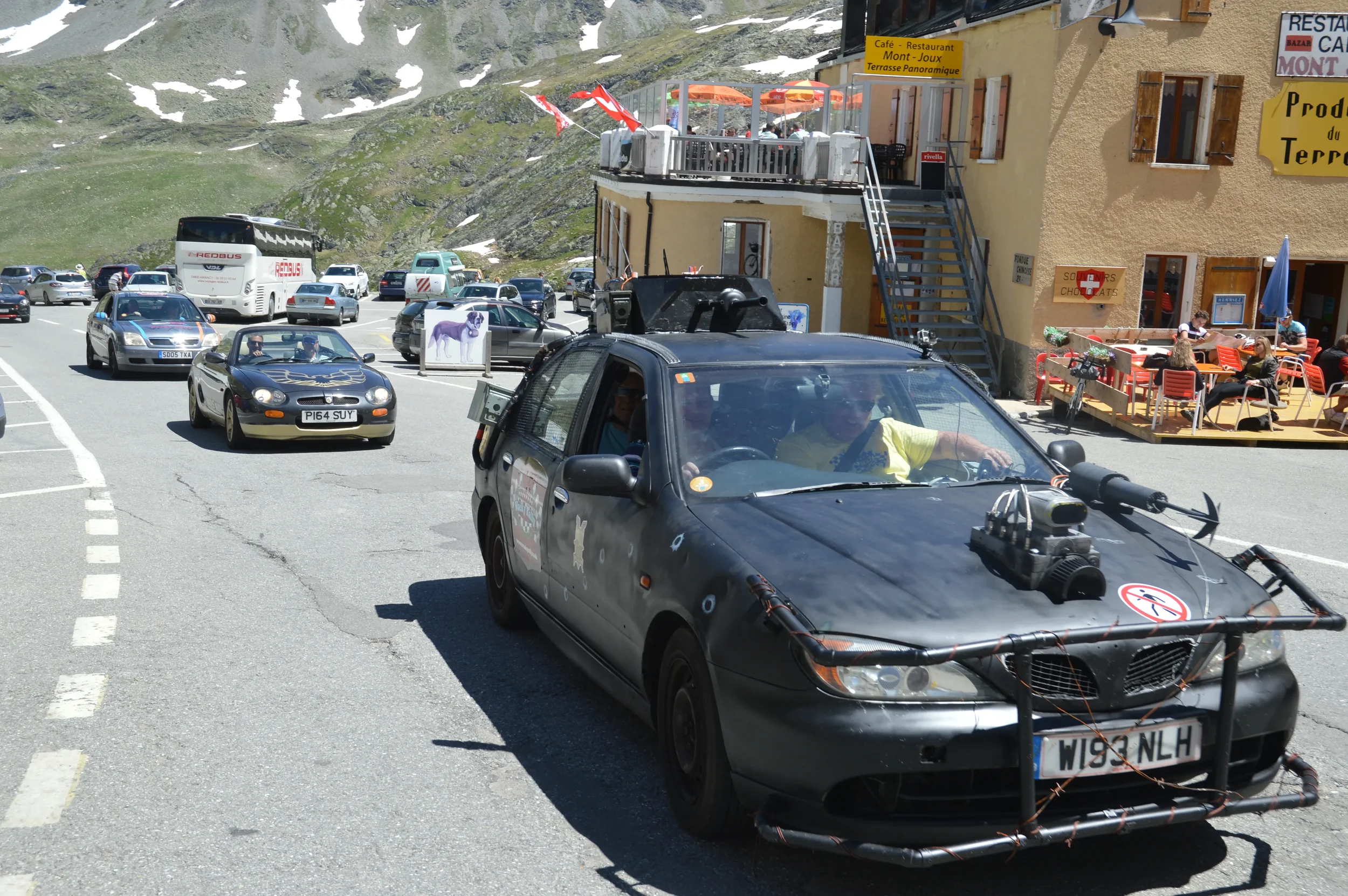 A black car with a mounted camera rig driving in a mountainous area with many parked cars and a restaurant with outdoor seating.