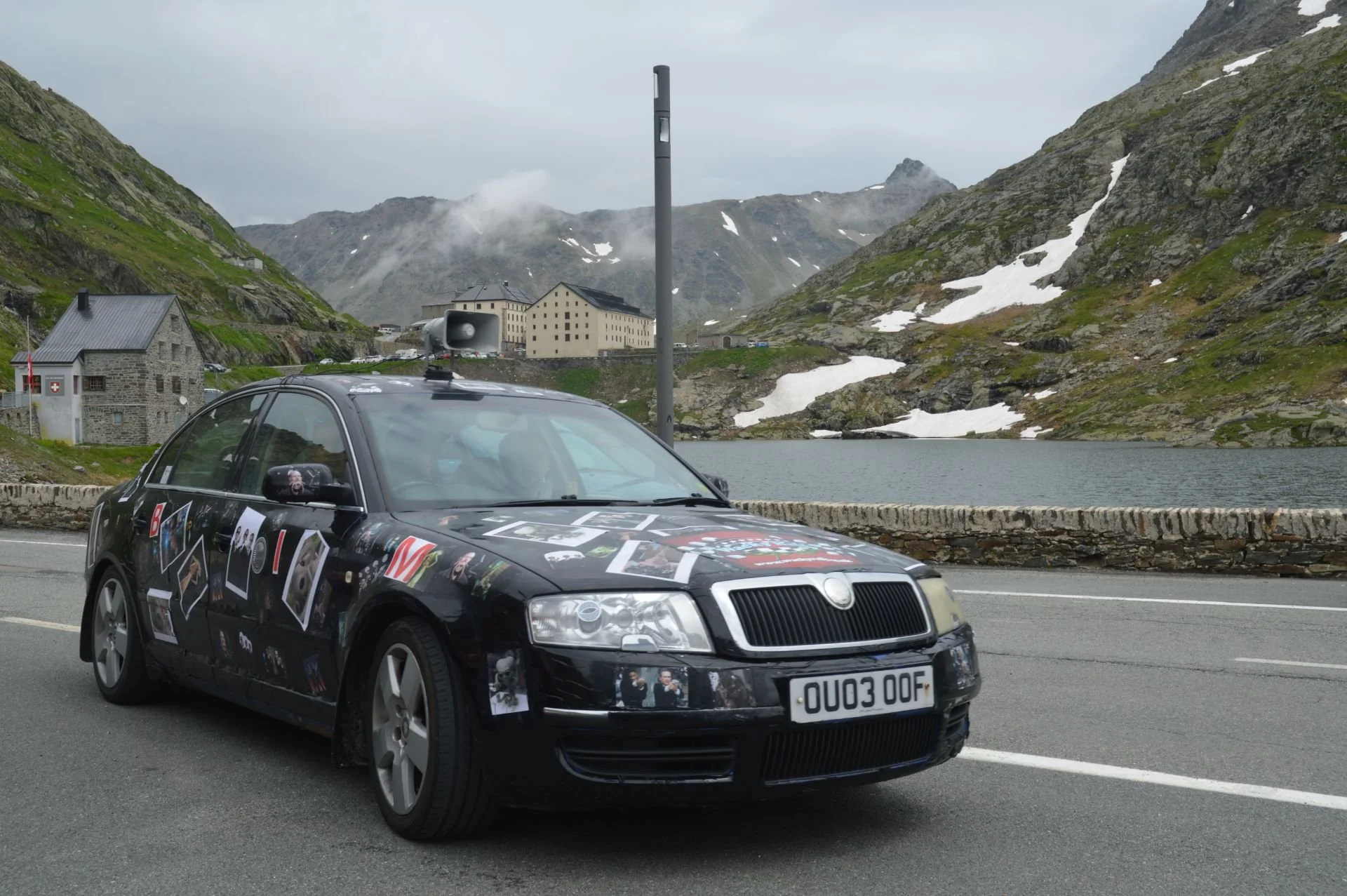 A black car decorated with various pictures and stickers on its body, driving on a mountain road near a lake, with snow-capped mountains and buildings in the background.