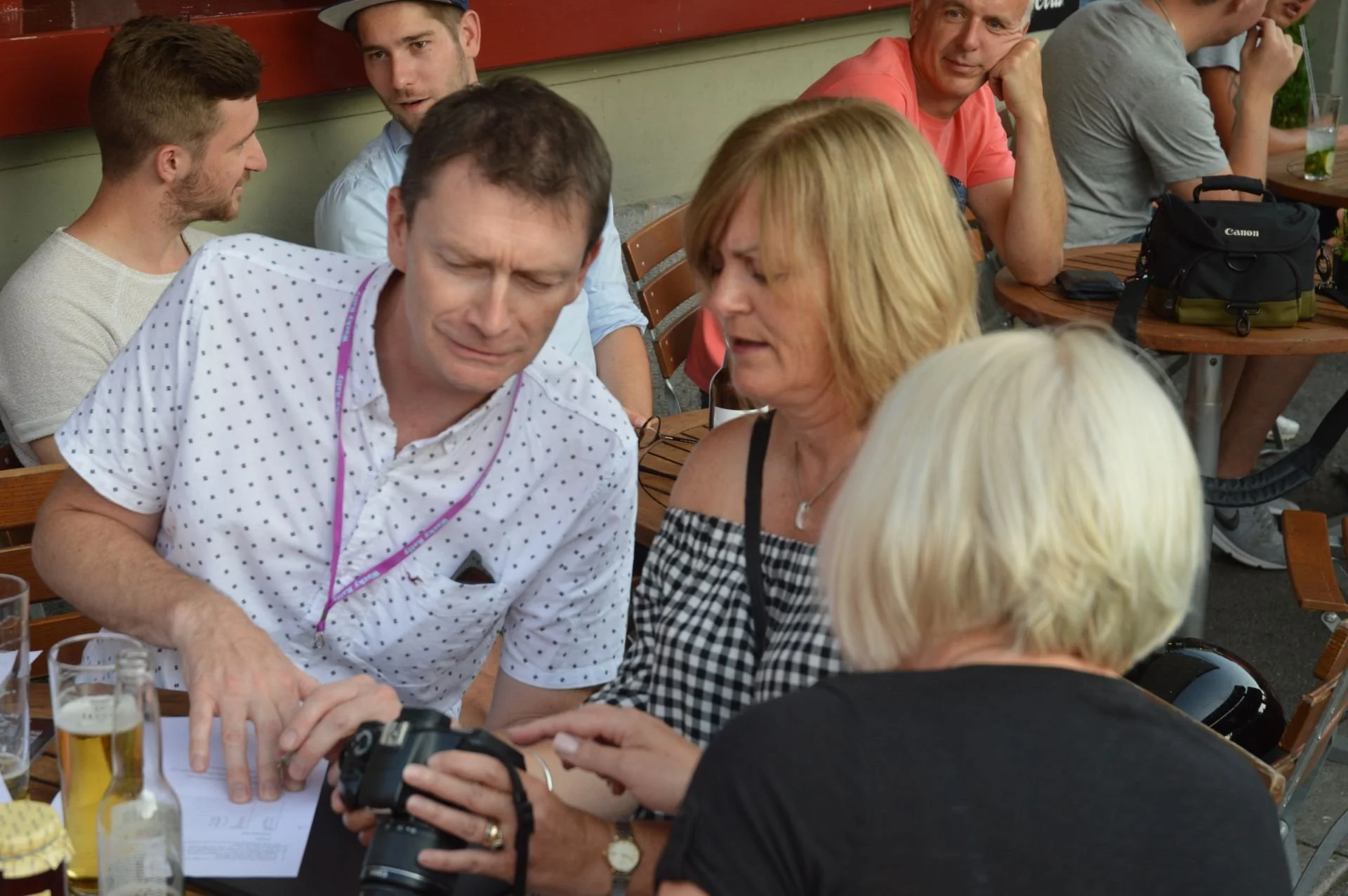 People seated around a table at an outdoor cafe, with one person showing a camera to others while looking at a piece of paper.