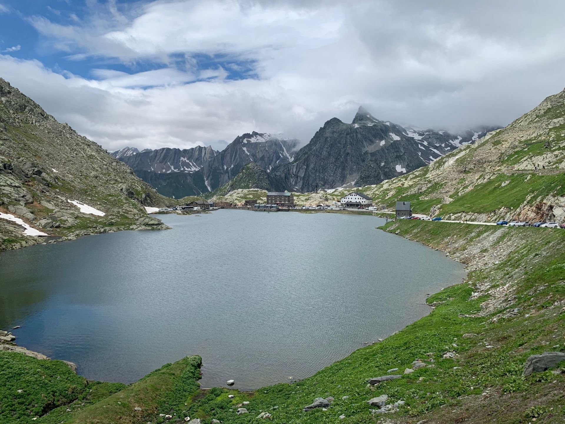 A mountain lake surrounded by green grass and rocky slopes, with snow-capped peaks in the background and a small village with buildings near the water.