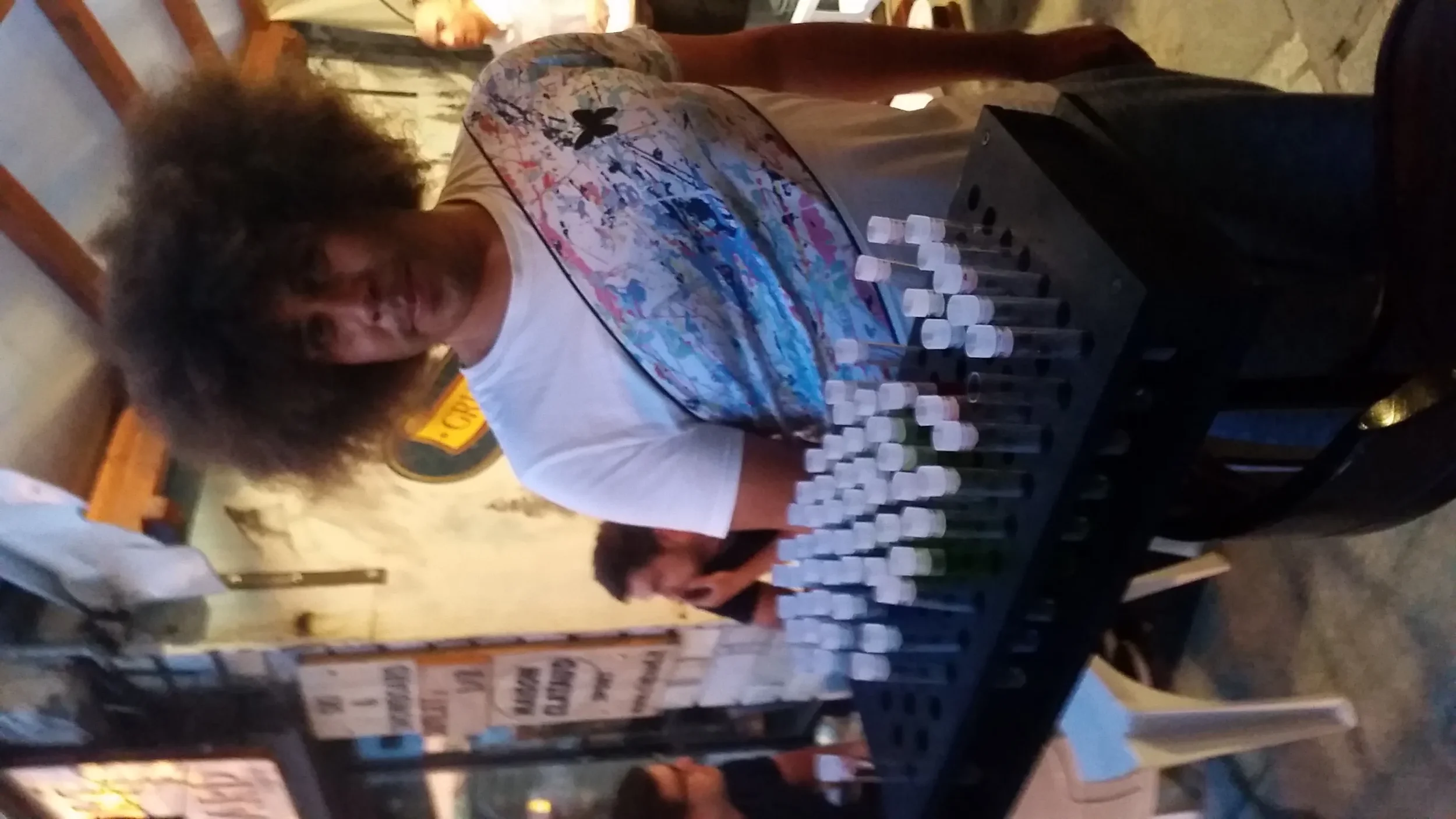 A person with voluminous curly hair standing behind a table covered with test tubes. They are in a casual setting with wooden chairs and some signs or menus in the background.