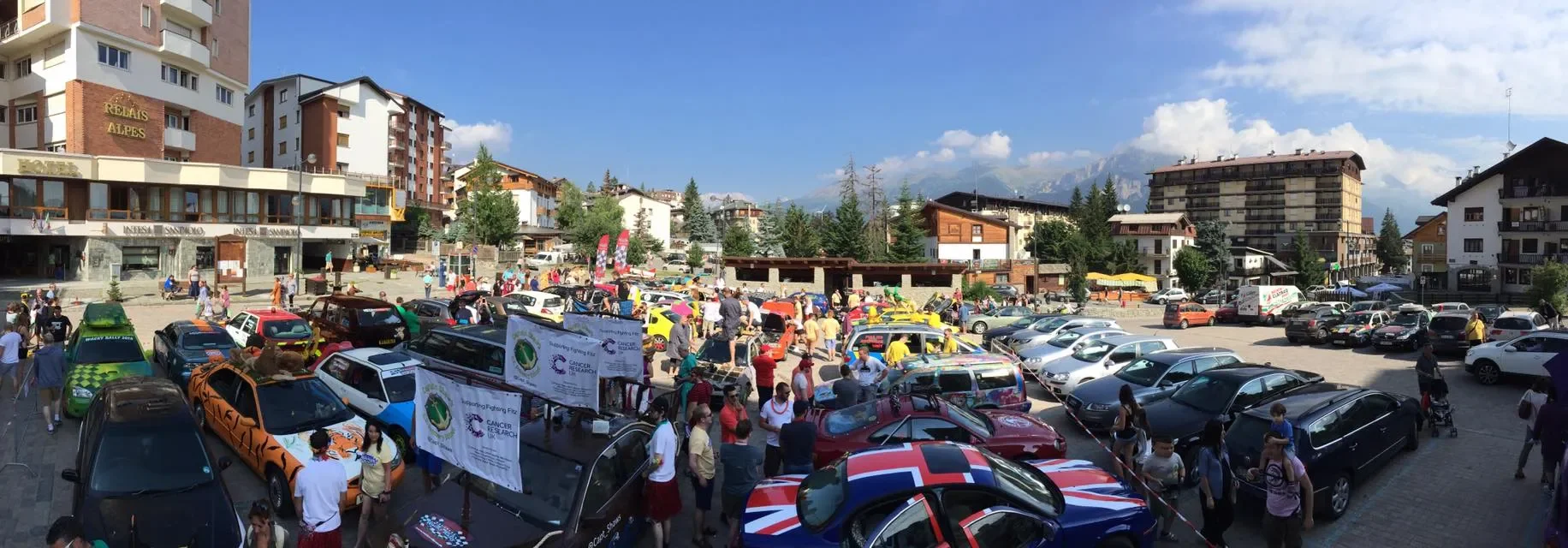 A busy outdoor car show in a parking lot with numerous colorful cars, including some with patriotic and themed designs, surrounded by many people.