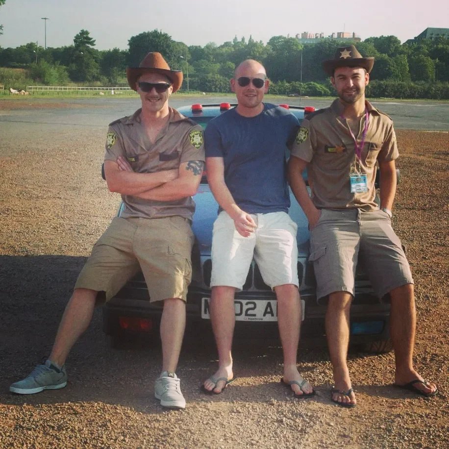 Three men in casual summer clothes and sunglasses, two dressed as police officers with sheriff hats, are sitting on the hood of a police car in an outdoor parking lot during daytime.