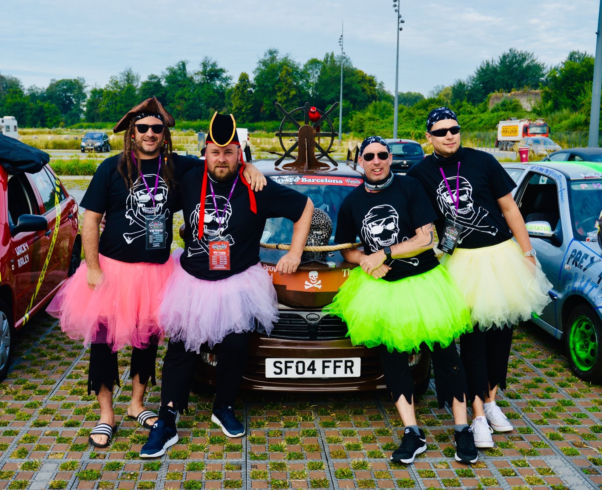 Four men in pirate costumes with tutus standing in front of a decorated car at a parking lot.
