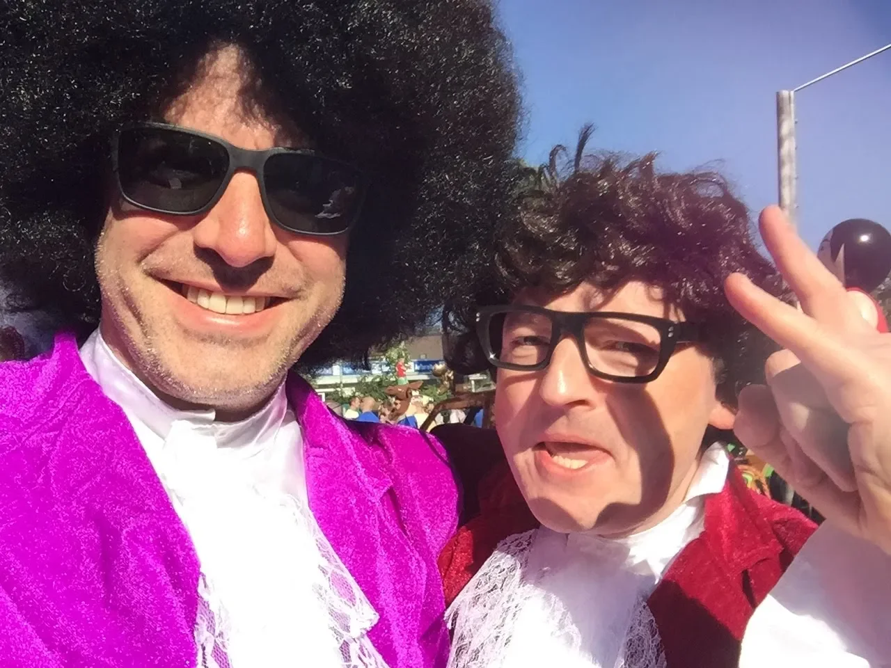 Two men dressed as Elvis Presley with black wigs, glasses, and colorful jackets, smiling and taking a selfie outdoors.