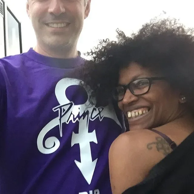 Smiling man wearing a purple Prince t-shirt and smiling woman with glasses and curly hair taking a selfie together.