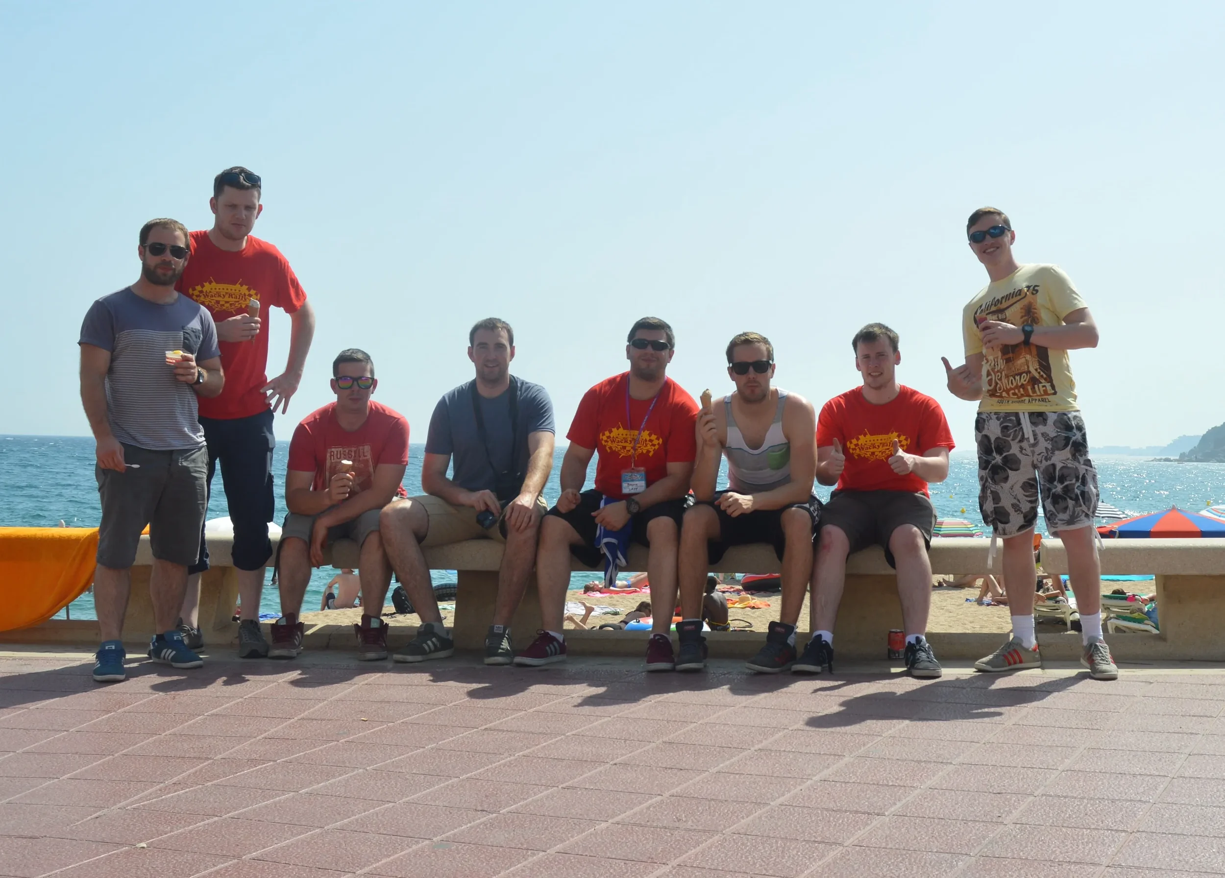 Group of ten young men at the beach, some sitting on a bench and others standing, with the ocean and colorful umbrellas in the background.