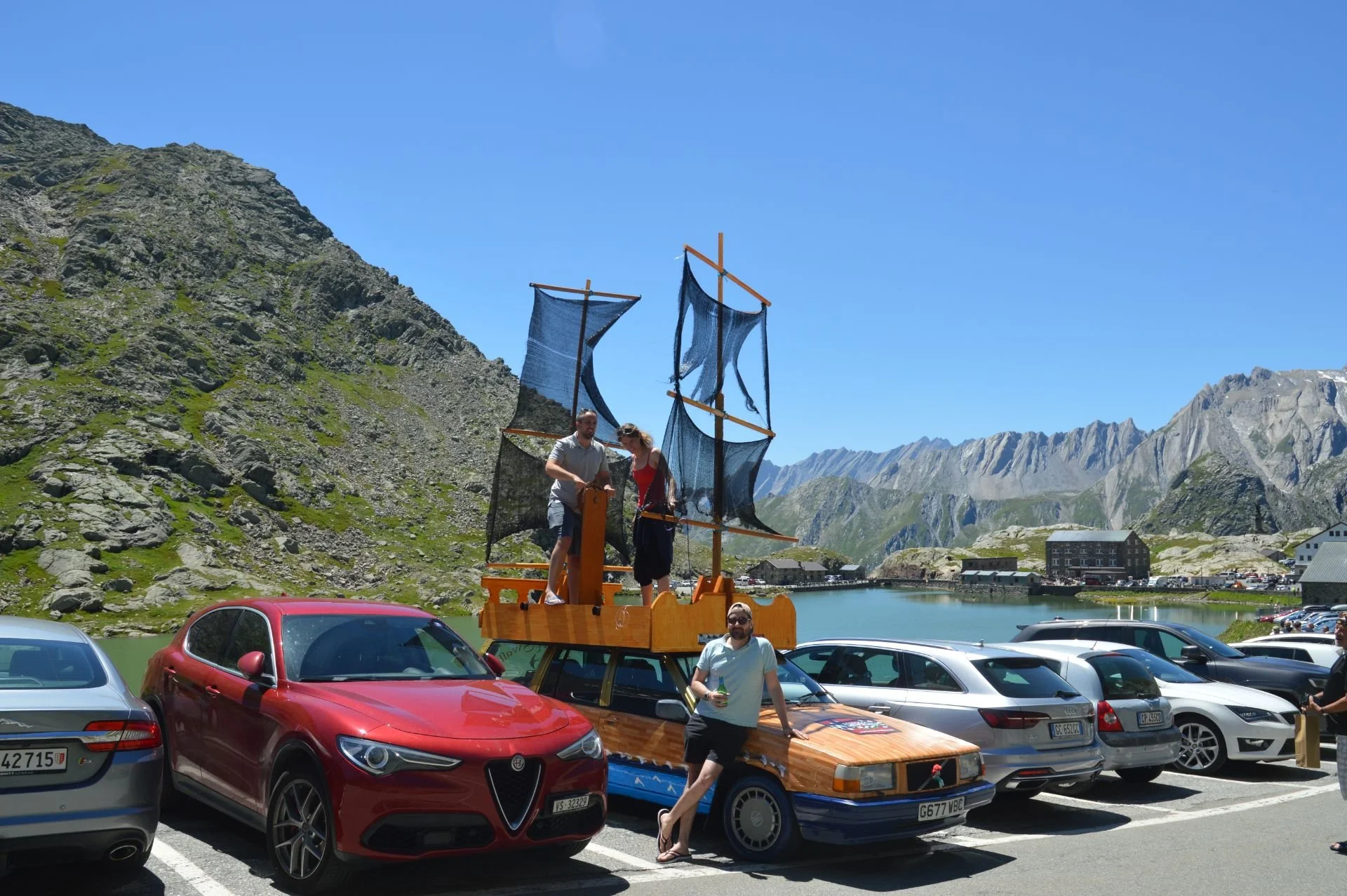 Several cars parked near a mountain lake with a decorative ship on top of a car, and people standing around it, in a scenic mountain landscape.
