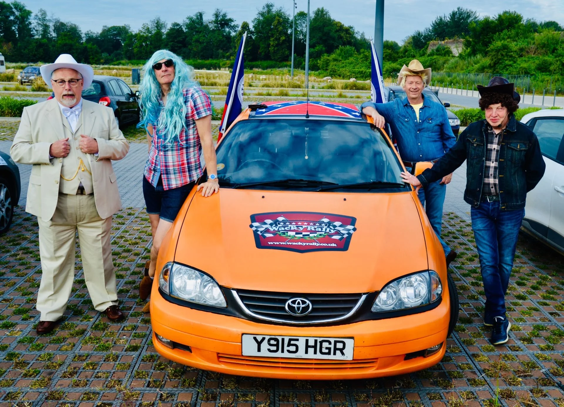 Group of five people in costumes posing around an orange Toyota car with a racing sticker on the hood and a UK license plate reading Y915 HGR, parked on a paved lot with greenery in the background.