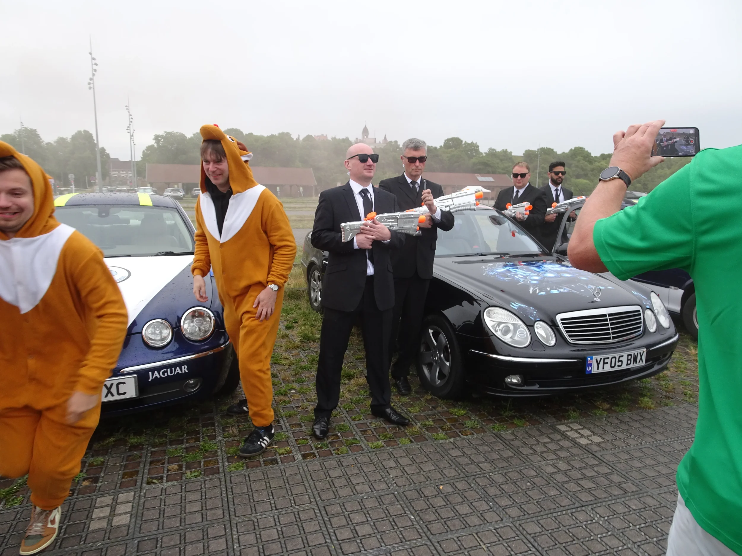 People dressed in animal costumes and suits holding toy water guns, standing in front of luxury cars, with one person recording the scene on their phone.
