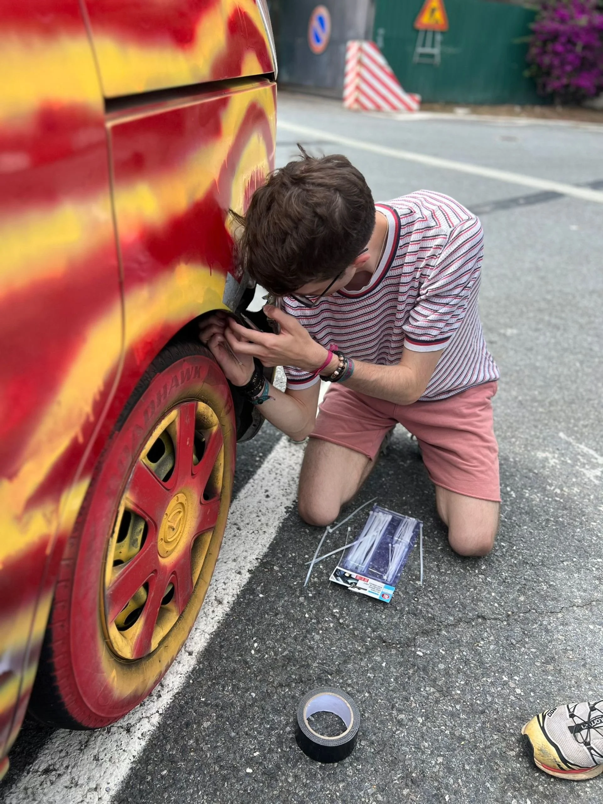 A person kneeling on the ground, working on the tire of a red and yellow painted car, with tools and tape nearby in an outdoor parking area.
