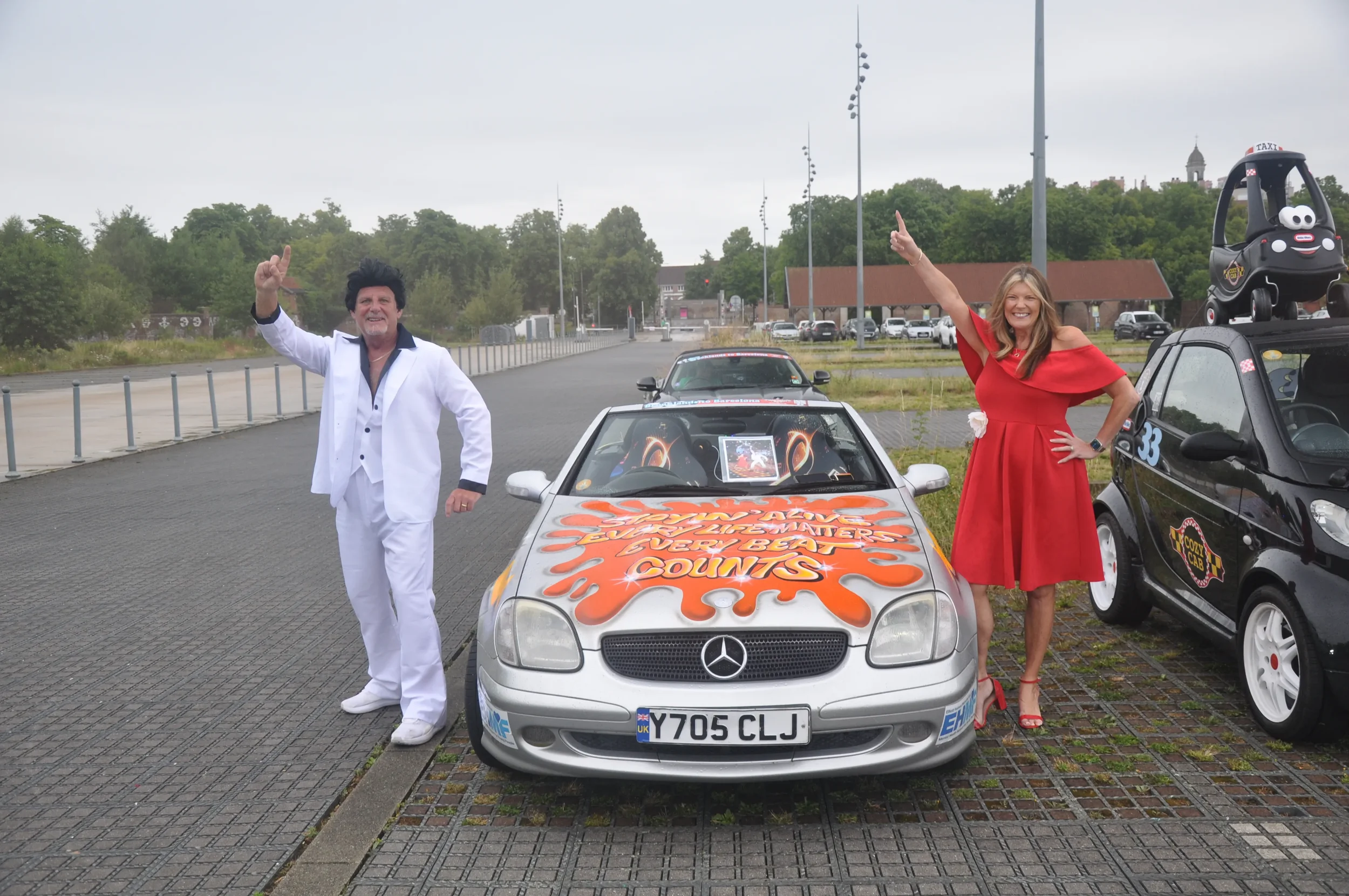 A man dressed as Elvis Presley in a white suit and woman in a red dress standing beside a colorful decorated Mercedes-Benz car, both smiling and pointing upwards, outdoors in a parking lot with trees and overcast sky.