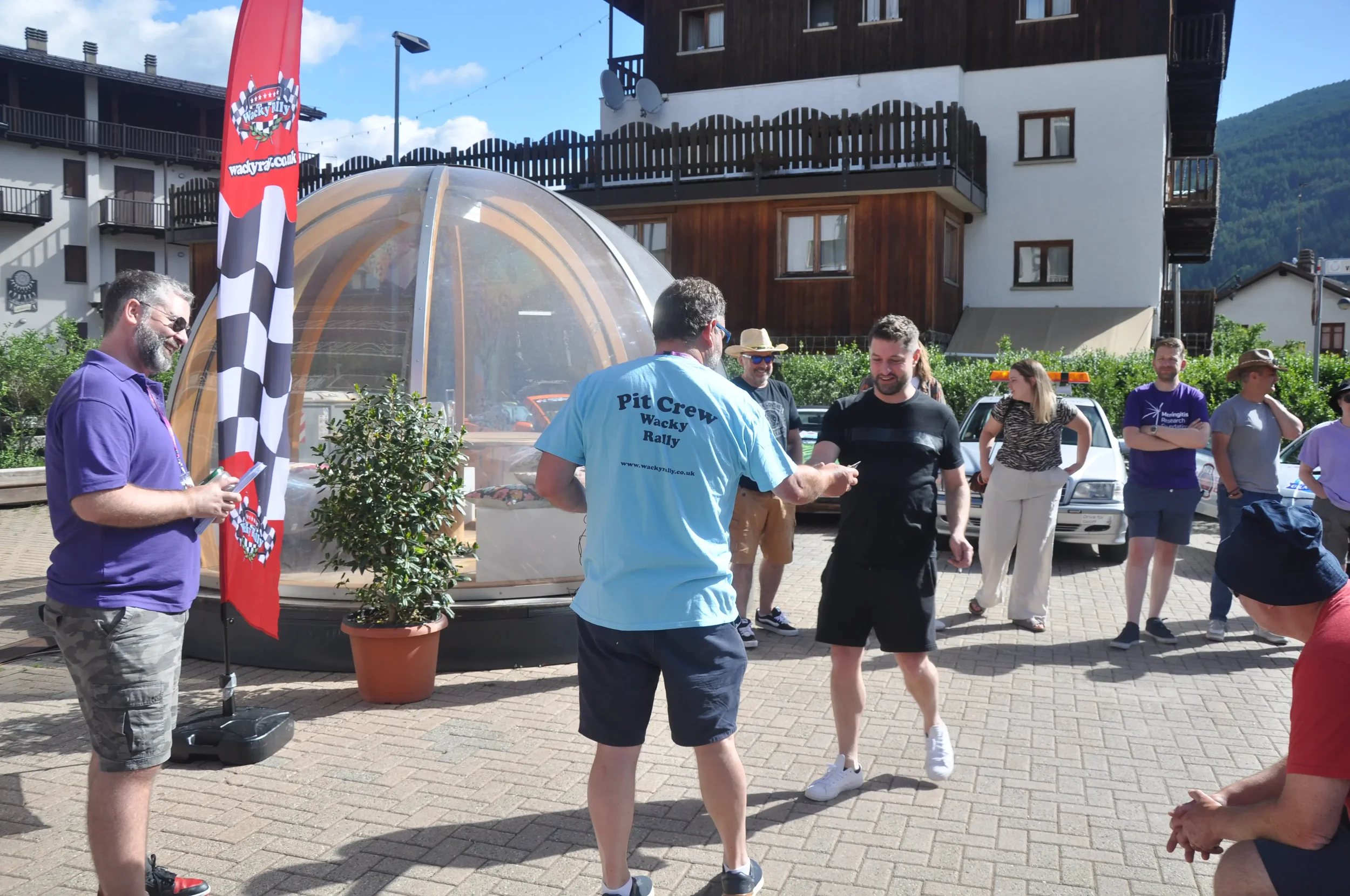 People gathered outdoors during a rally, with a man wearing a blue T-shirt reading 'Pit Crew Wacky Rally' handing something to another man. Others stand nearby, some smiling, with a small indoorstructure and a mountain in the background.