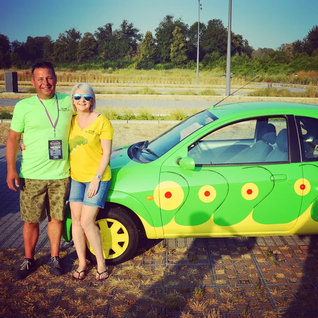 A man and woman standing side by side outdoors, smiling, near a small green car with a colorful, whimsical design resembling a peacock or other bird, in a parking lot with trees in the background.