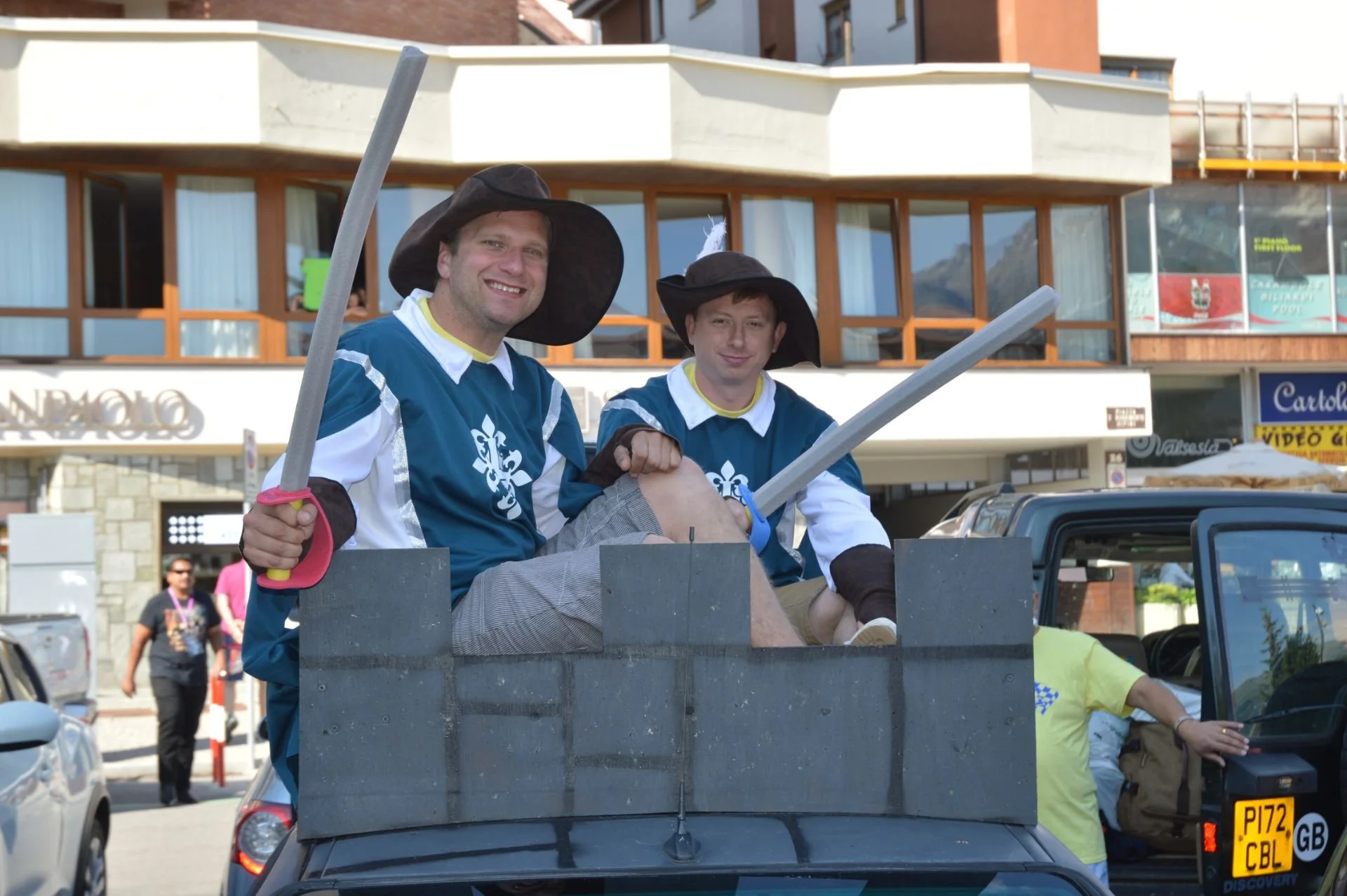 Two men dressed as medieval knights with swords, sitting in a forklift decorated to look like a castle, smiling at the camera during a festival or parade.