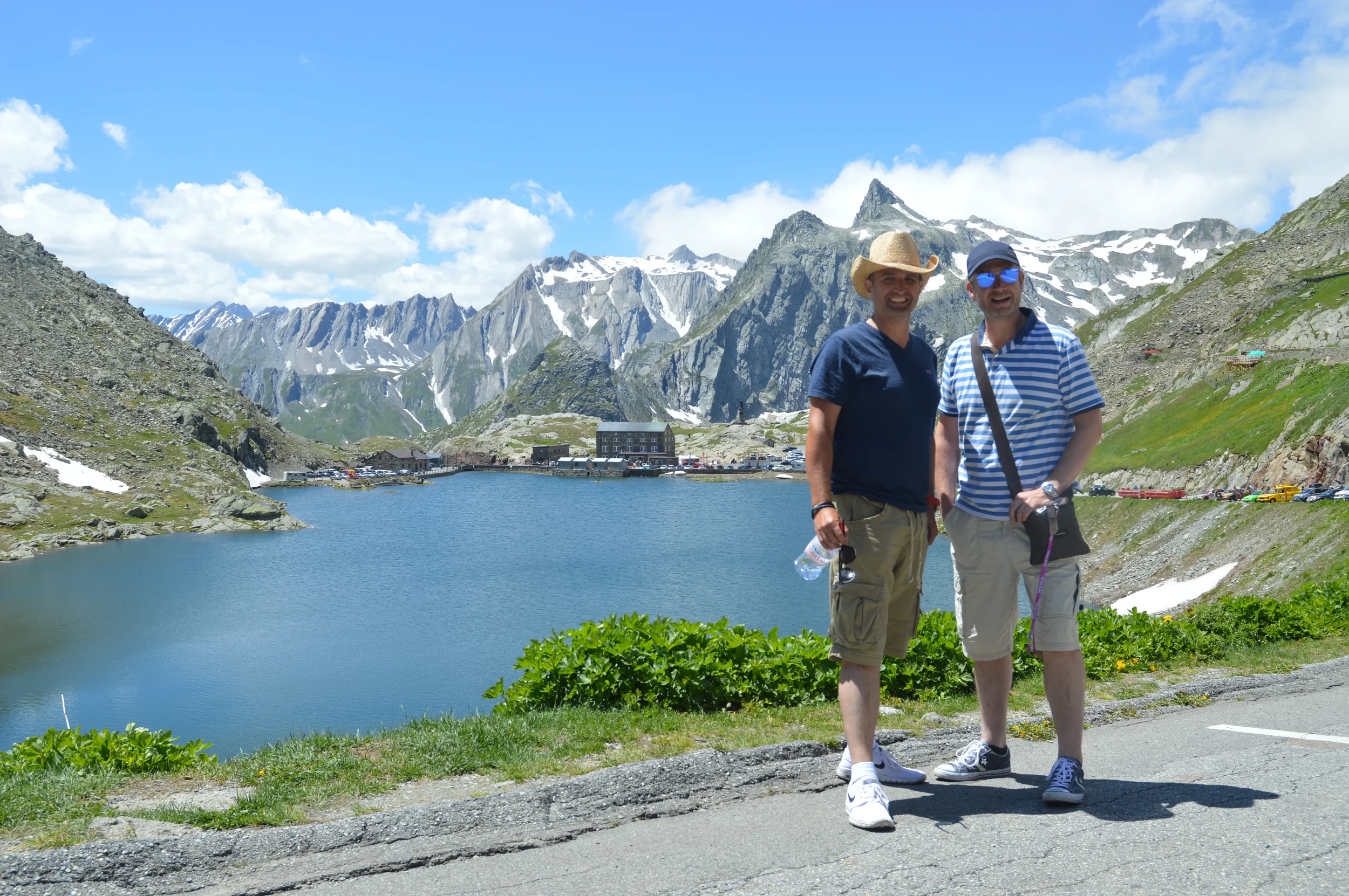 Two men standing on a pathway near a lake surrounded by mountains, with some snow patches and a partly cloudy sky, smiling at the camera.