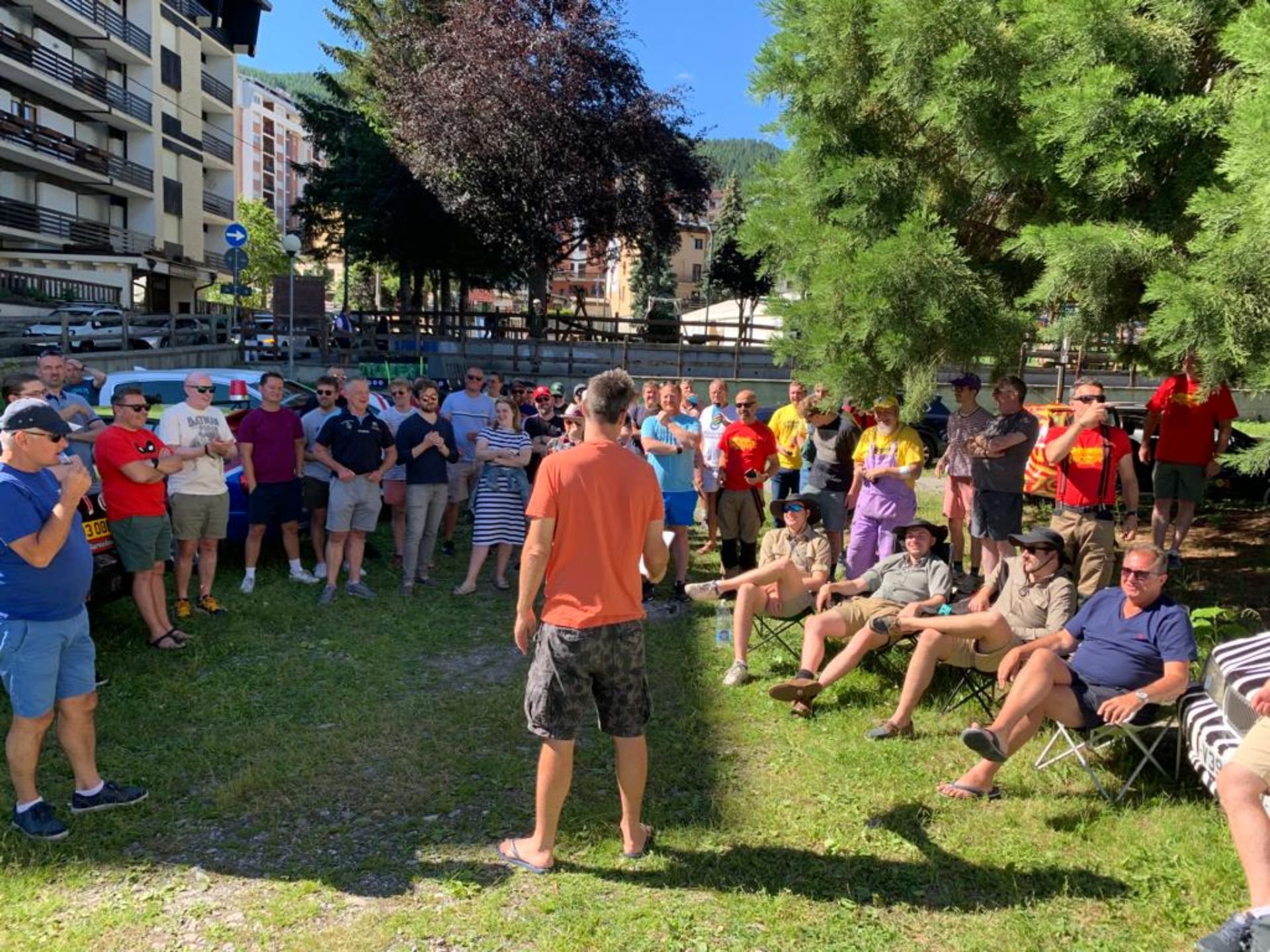 Group of people gathered outdoors in a park-like area, some sitting on chairs and others standing, listening to a speaker in an orange shirt. There are trees providing shade, and buildings and parked cars in the background.