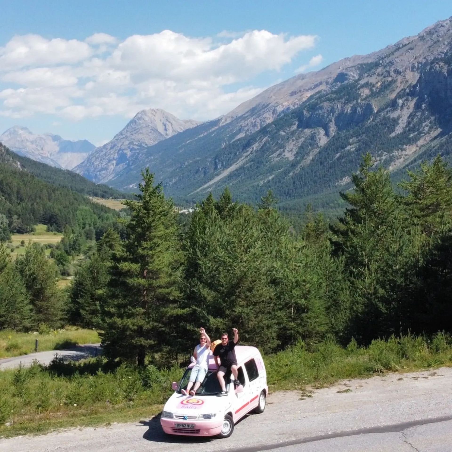Two people sitting on top of a small pink and white ice cream truck parked on the side of a road in a mountainous forested area with trees and mountains in the background.