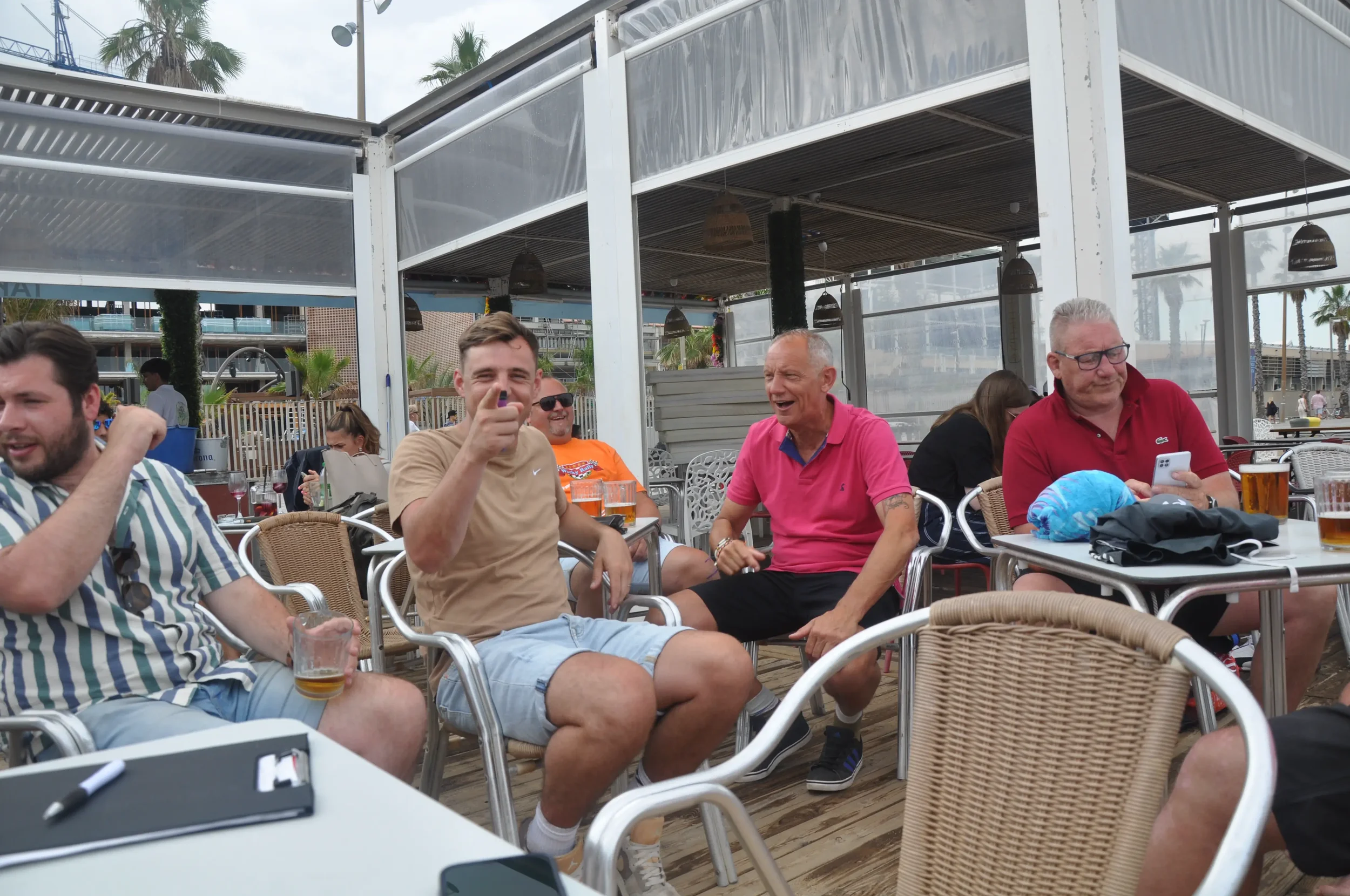 Group of people sitting at an outdoor bar or restaurant, laughing and socializing, with palm trees in the background.