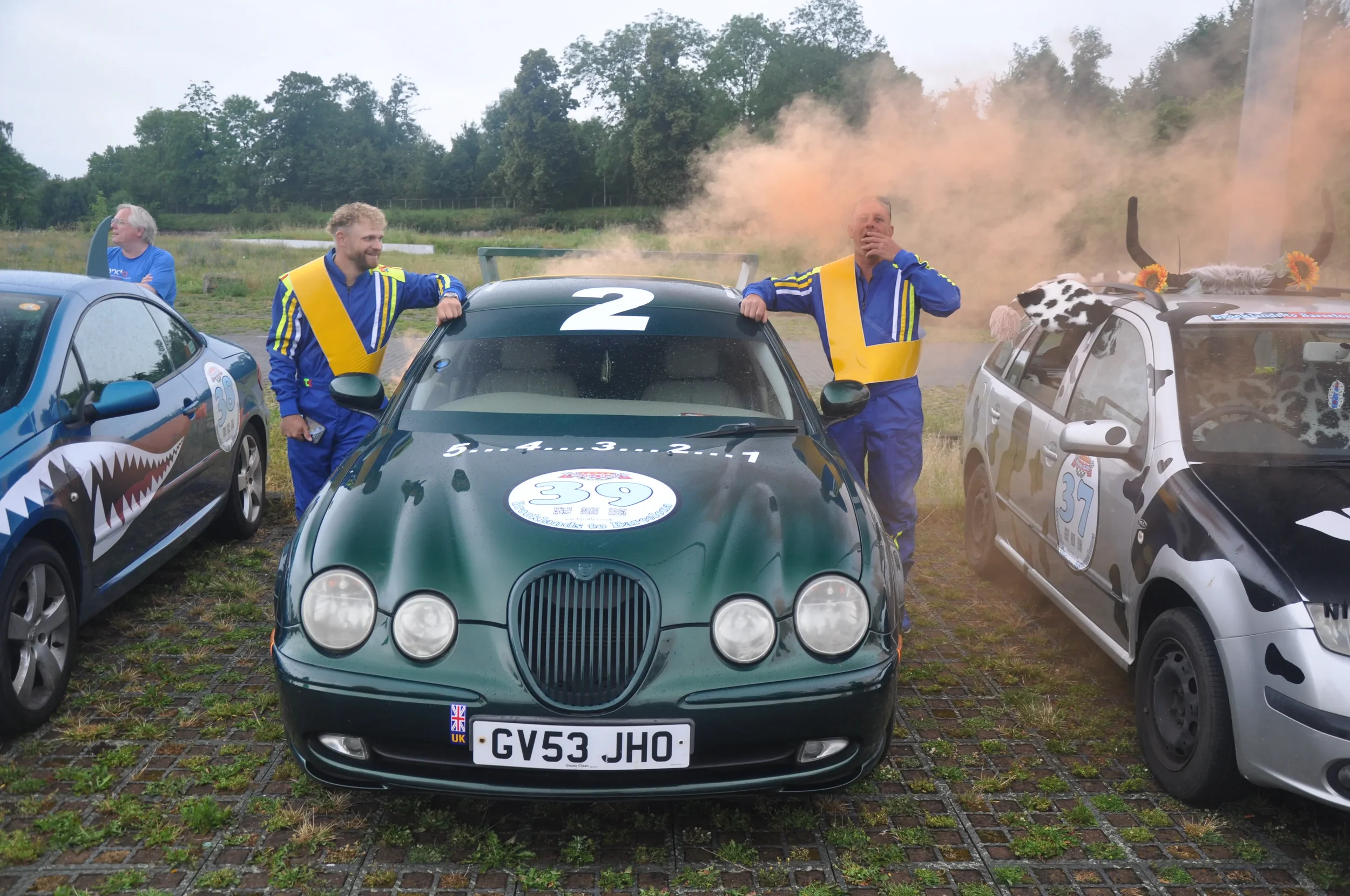 Three cars parked on a gravel lot with three men in racing suits standing next to them. One car has the number 39, another has the number 37, and the third has the number 2. The men are celebrating, with one man appearing surprised and another smilin