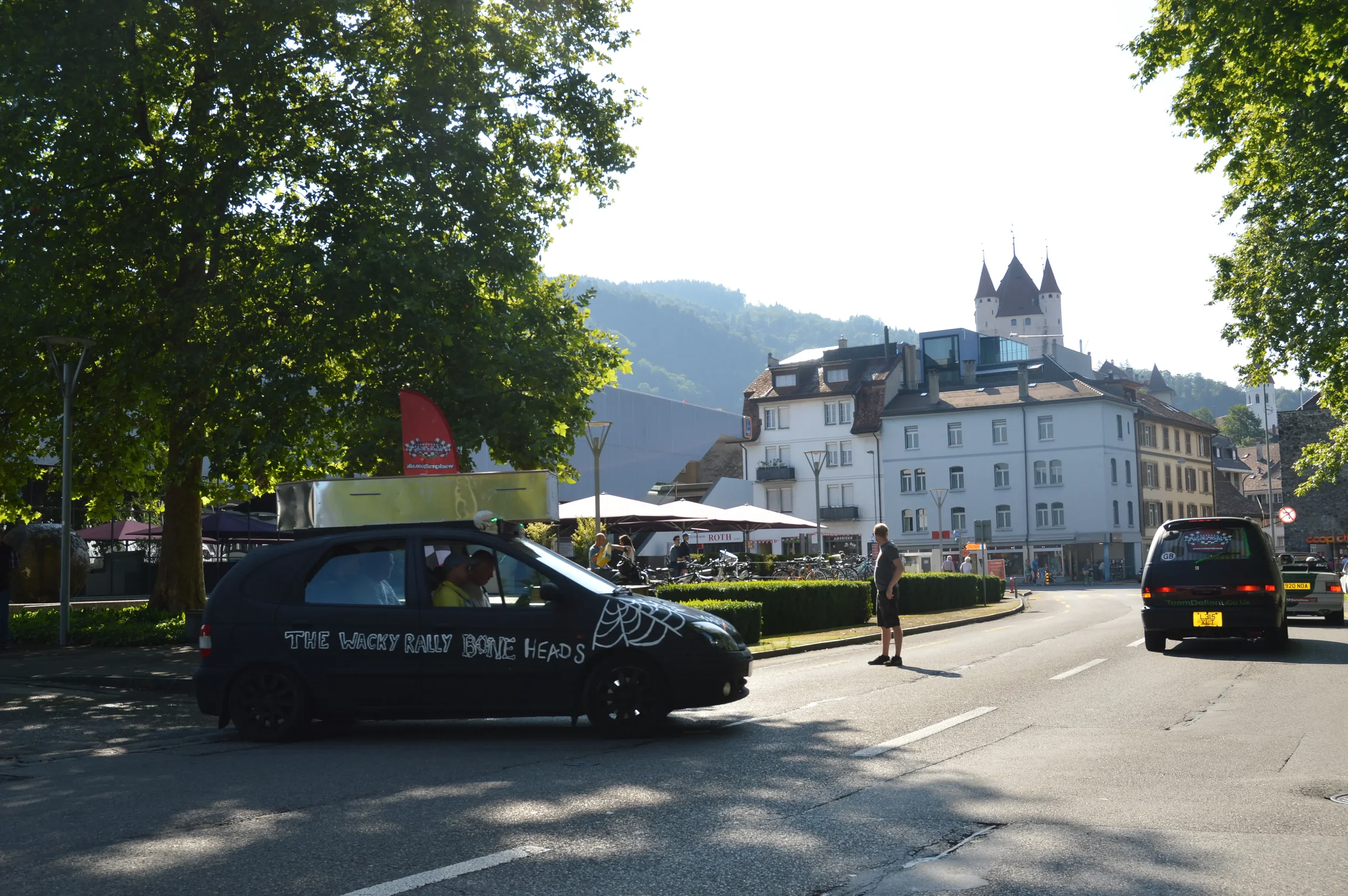 A small black car decorated with a spider web and the phrase 'THE WACKY RALLY BONE HEADS' on the side, parked on a city street. People are walking, and there are trees, white buildings, and a castle-like structure in the background.