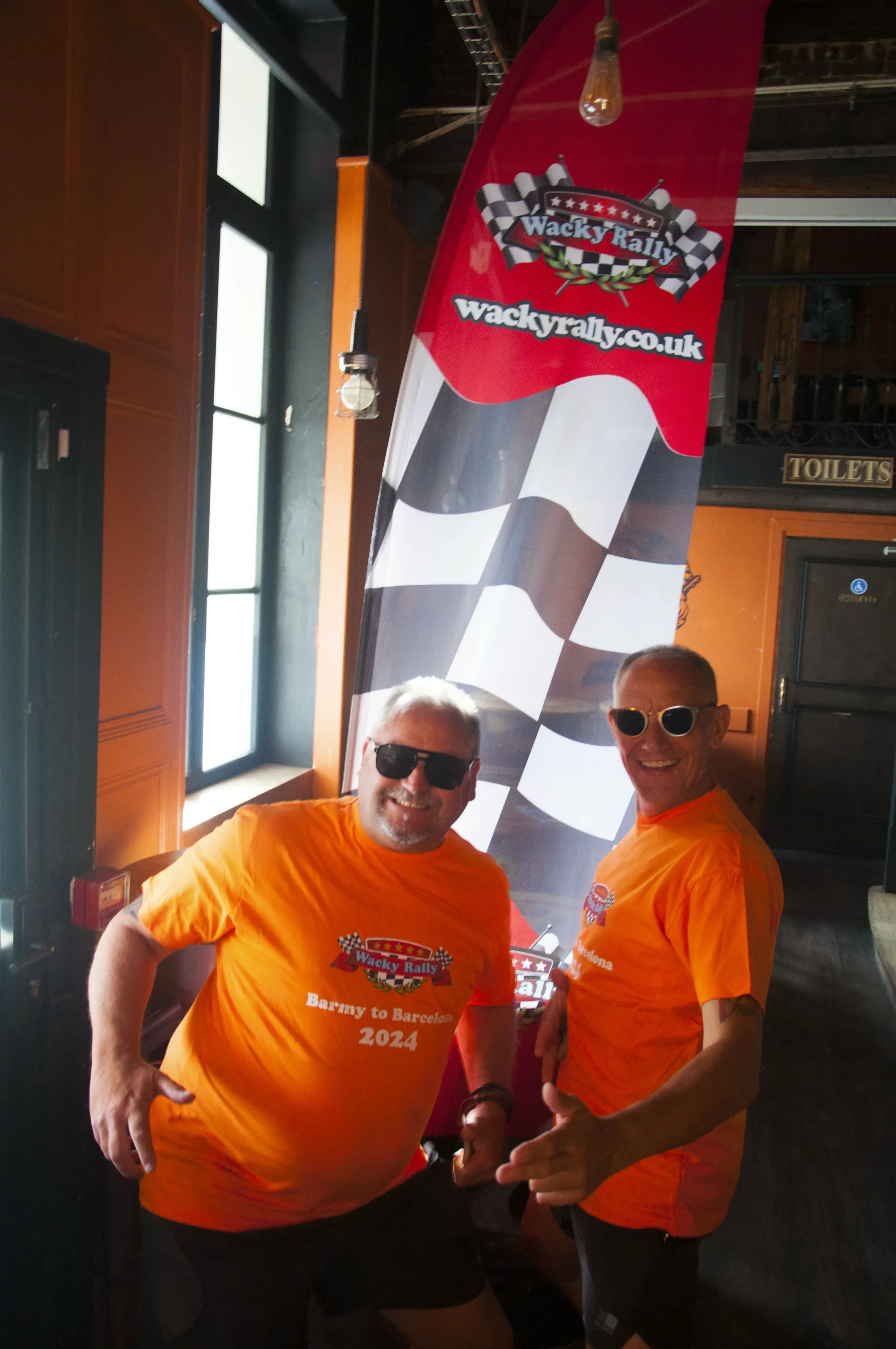 Two men in orange shirts and sunglasses smiling and posing for a photo in front of a large checkered flag banner with the text 'Wacky Rally' and website 'wackyrally.co.uk'.