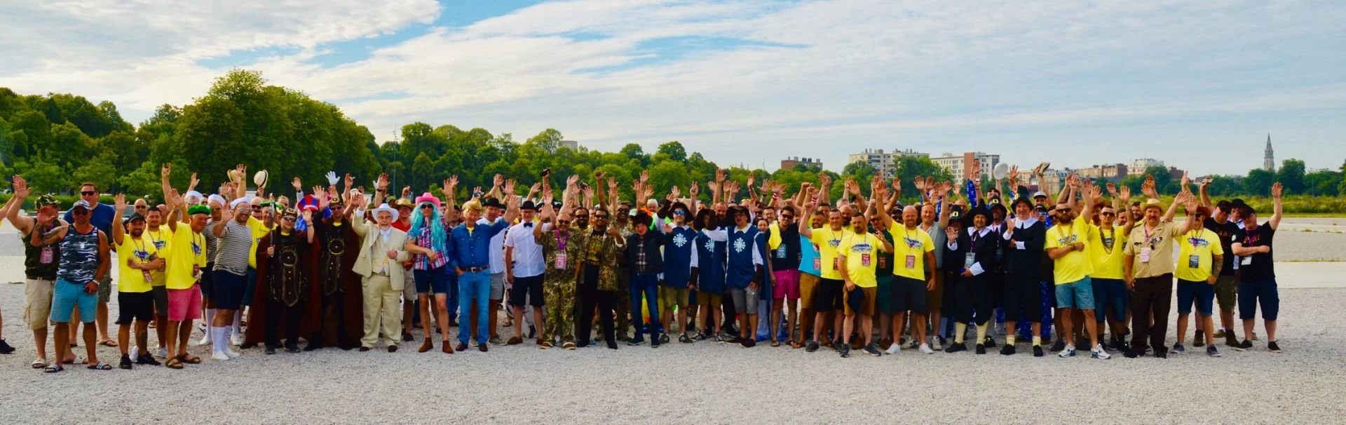 A large group of people posing outdoors on a gravel area, many with their hands raised, with trees and city buildings in the background, under a partly cloudy sky.