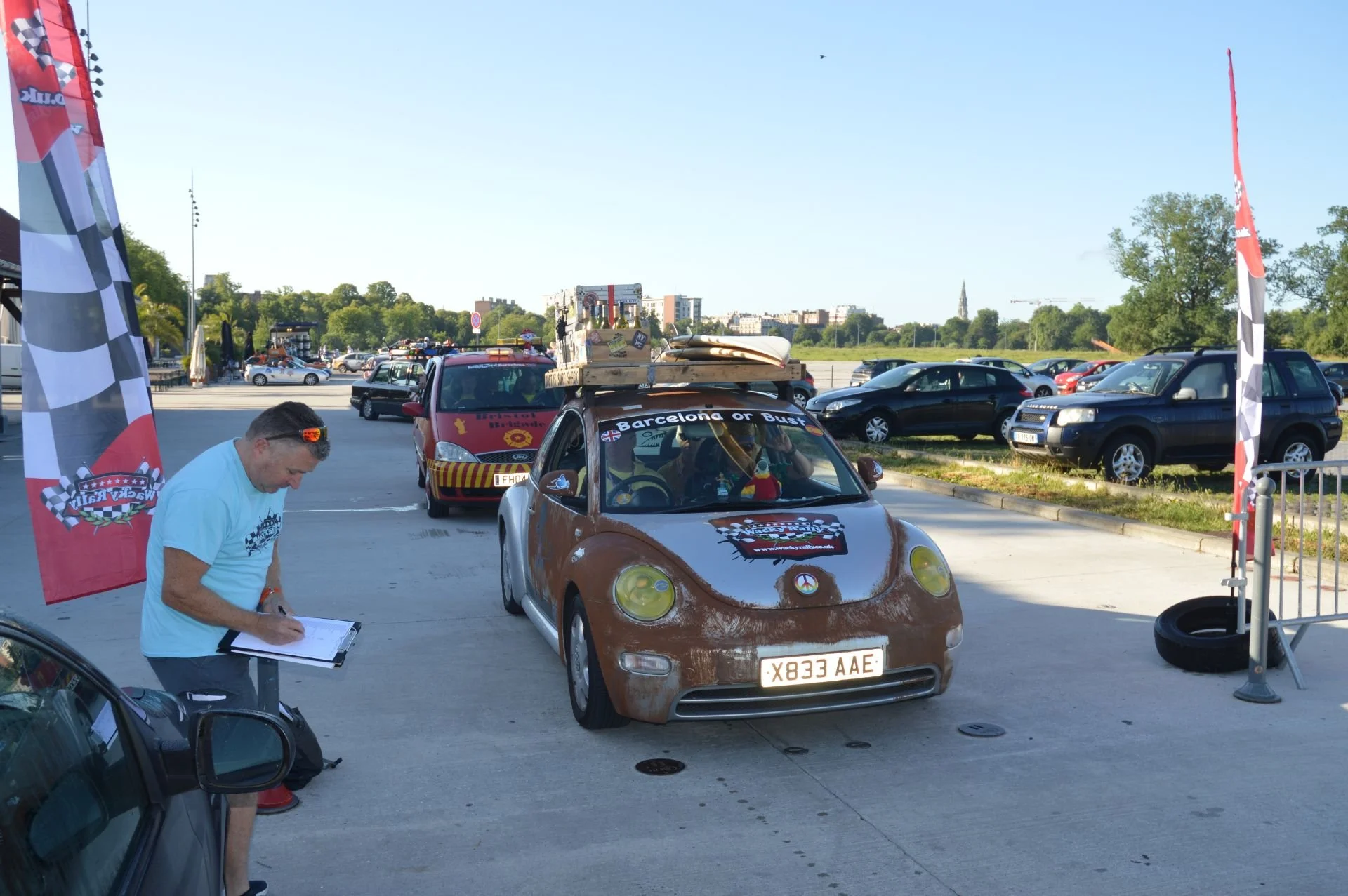 A brown car with a roof rack carrying a surfboard and boxes, parked near a racing event with banners that read 'Track Day' and 'Back in the Race.' A man in a light blue shirt is writing on a clipboard next to the car, with several other cars and tree