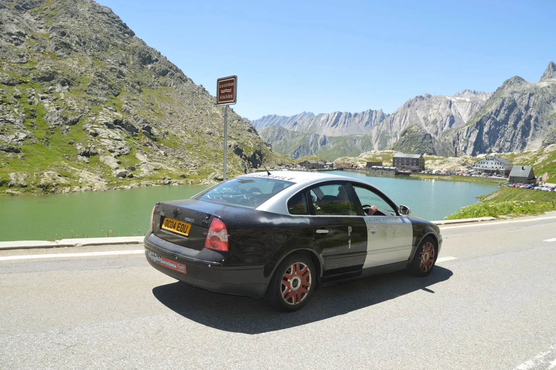 Police car parked by a mountain lake with green water, surrounded by rocky slopes and mountain peaks, under a clear blue sky.
