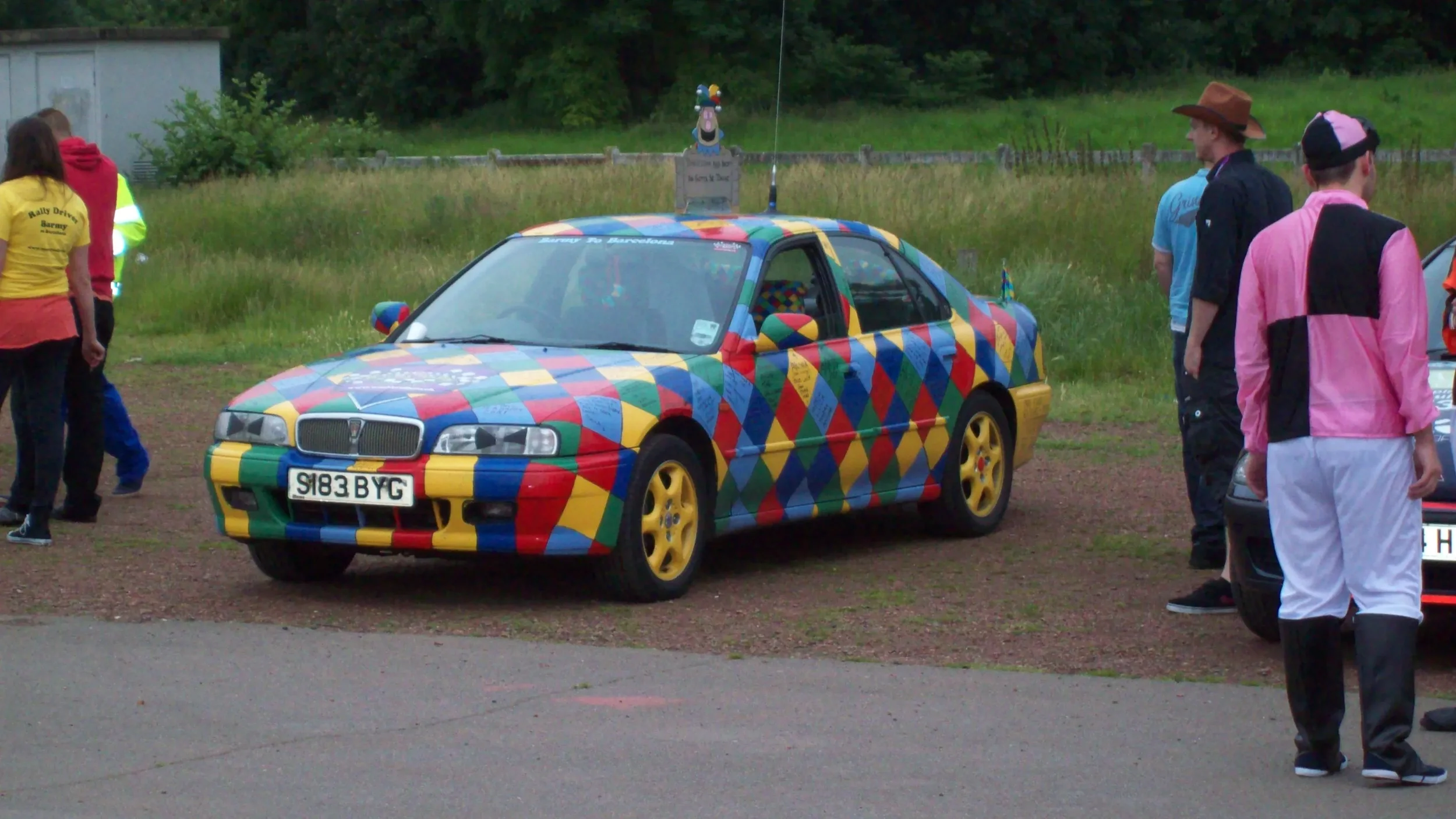 A car with a colorful diamond pattern wrap parked outdoors with several people around it, some in casual clothing and one in a pink jacket.