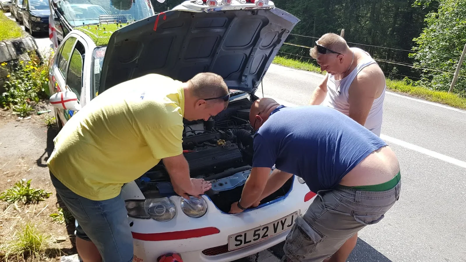 Four men work together to inspect or repair the engine of a white car with a Slovak license plate, parked on the side of a rural road on a sunny day.