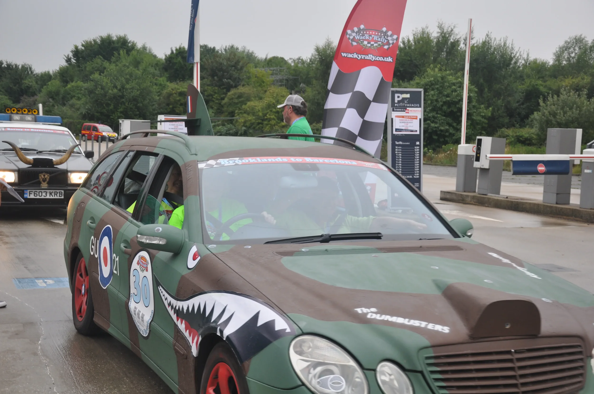 A green car with humorous shark design and race stickers, parked at a rally event, with a person inside wearing a neon yellow jacket and other rally cars and flags in the background.