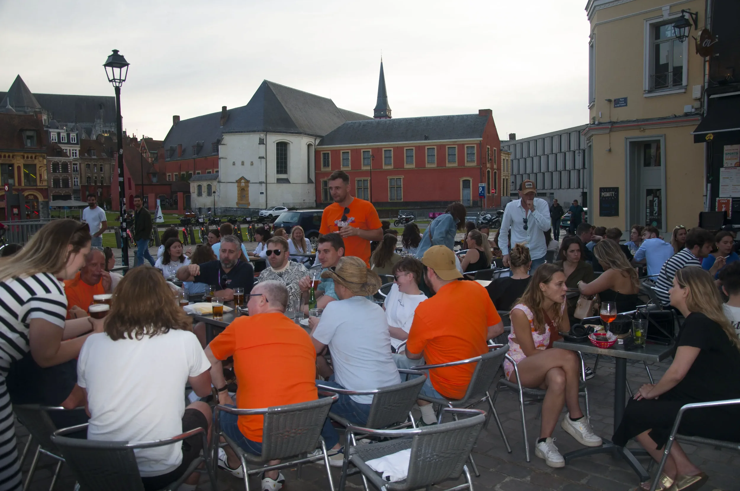 People sitting at outdoor cafe tables, socializing and drinking, with historic buildings and a church steeple in the background during evening.