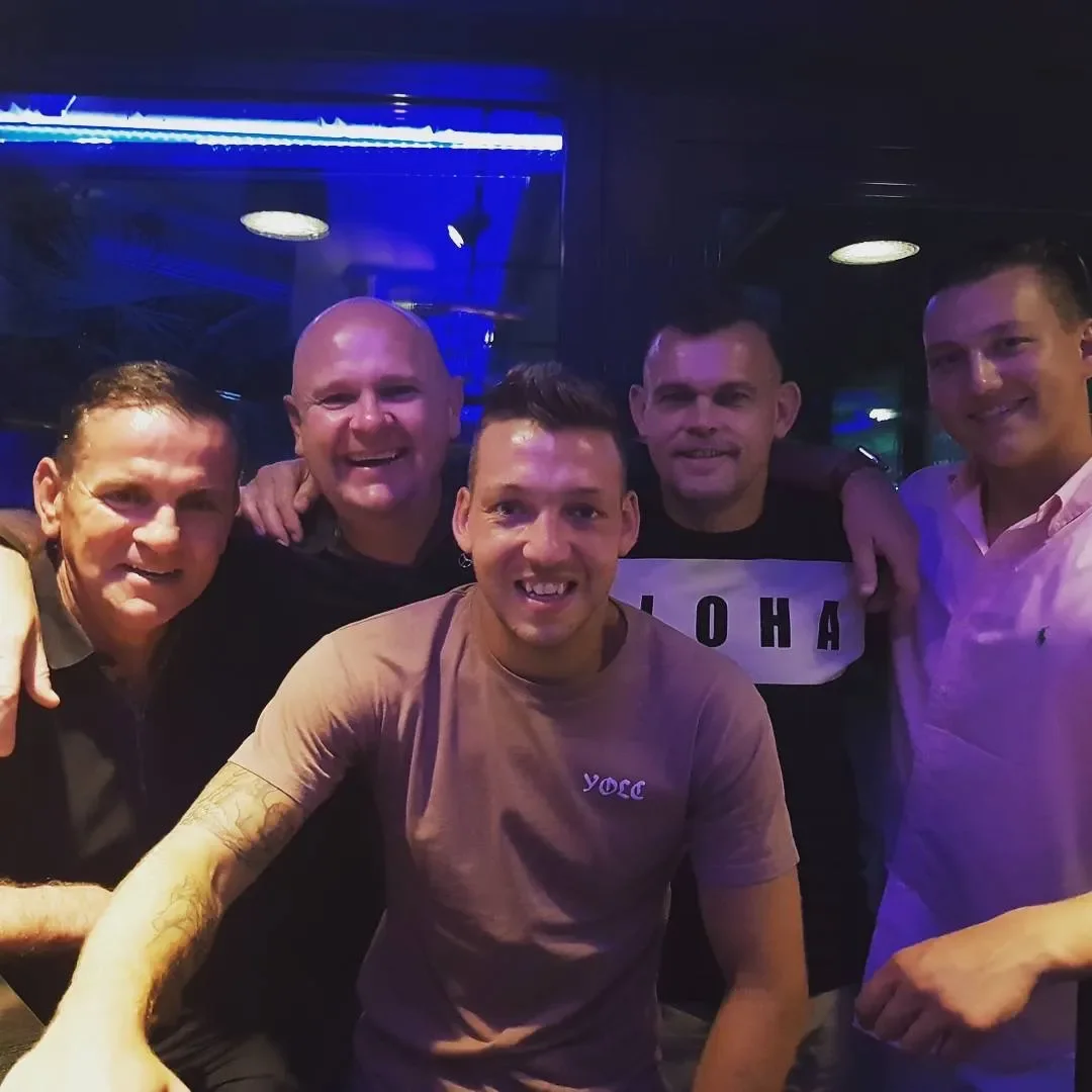 Group of six men smiling and posing together in a dimly lit nightclub or bar with blue lighting.