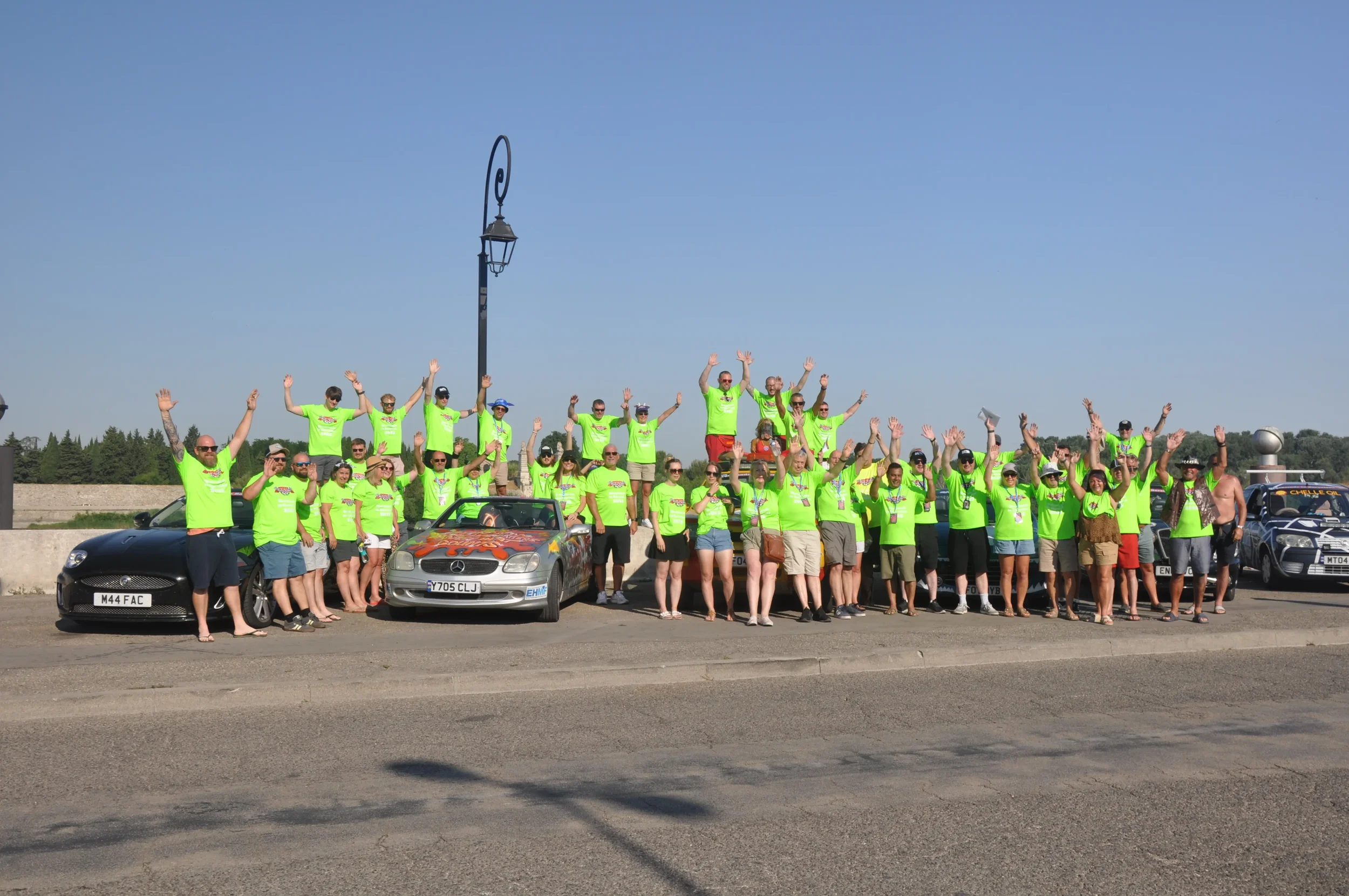 Large group of people wearing matching bright green shirts standing on a road with cars behind them, raising their hands in celebration, during a sunny day.