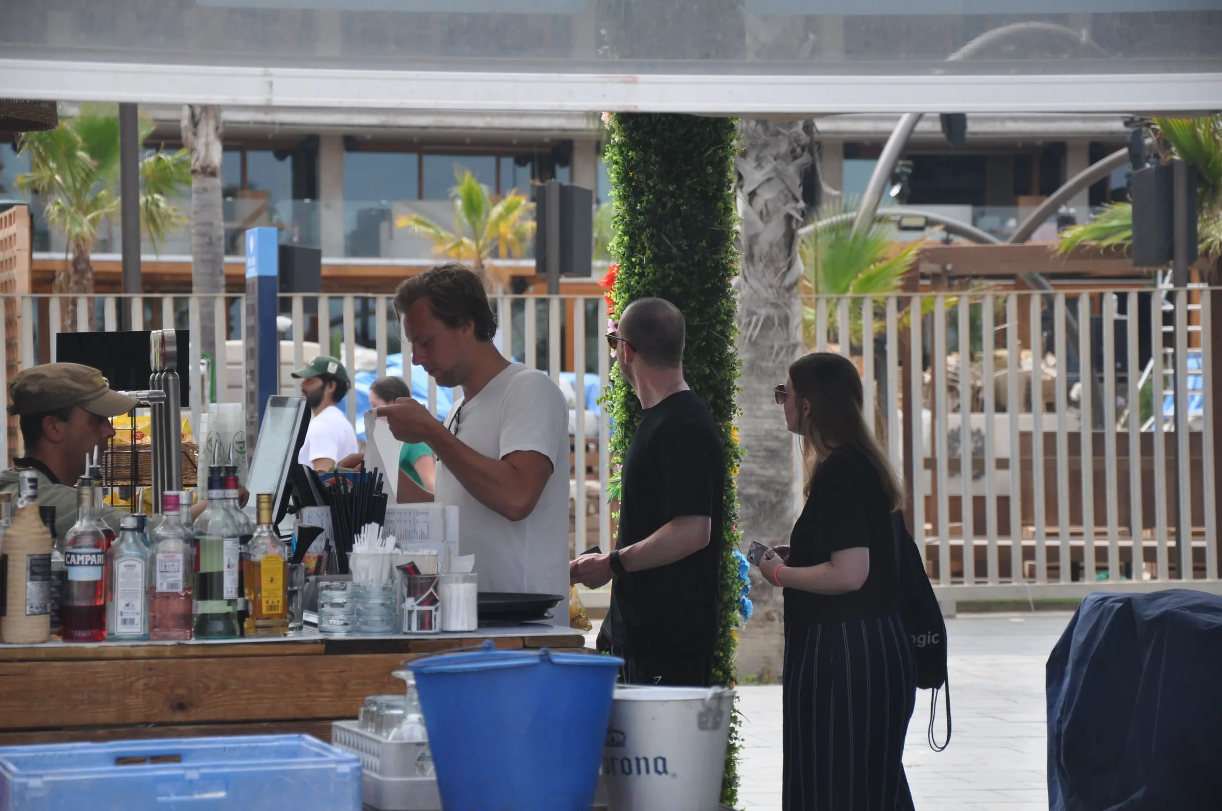 People standing at an outdoor bar by the pool, ordering drinks, with palm trees and pool chairs in the background.
