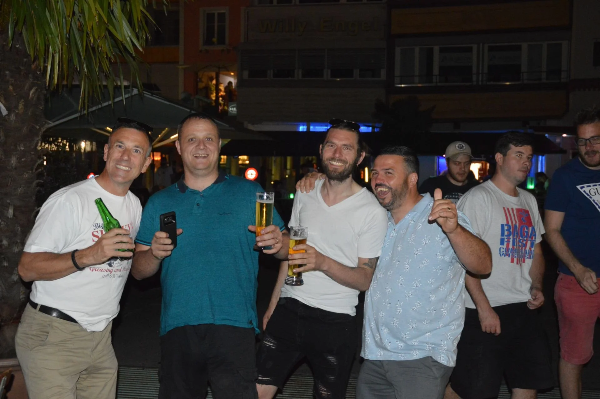 Group of five men smiling and holding drinks at a social gathering outdoors at night.