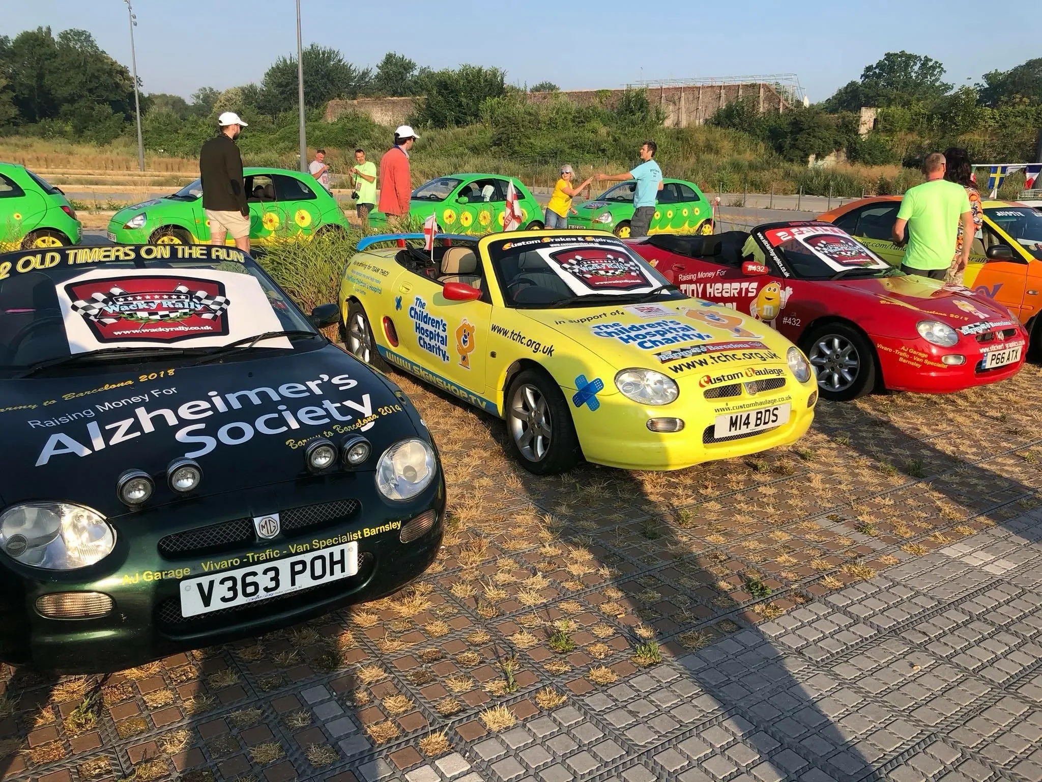 Multiple colorful cars parked outdoors at a charity event, with people gathering and walking around, and some cars decorated with promotional and charity logos, including Alzheimer's Society, Children’s Hospital Charity, and Tiny Hearts.