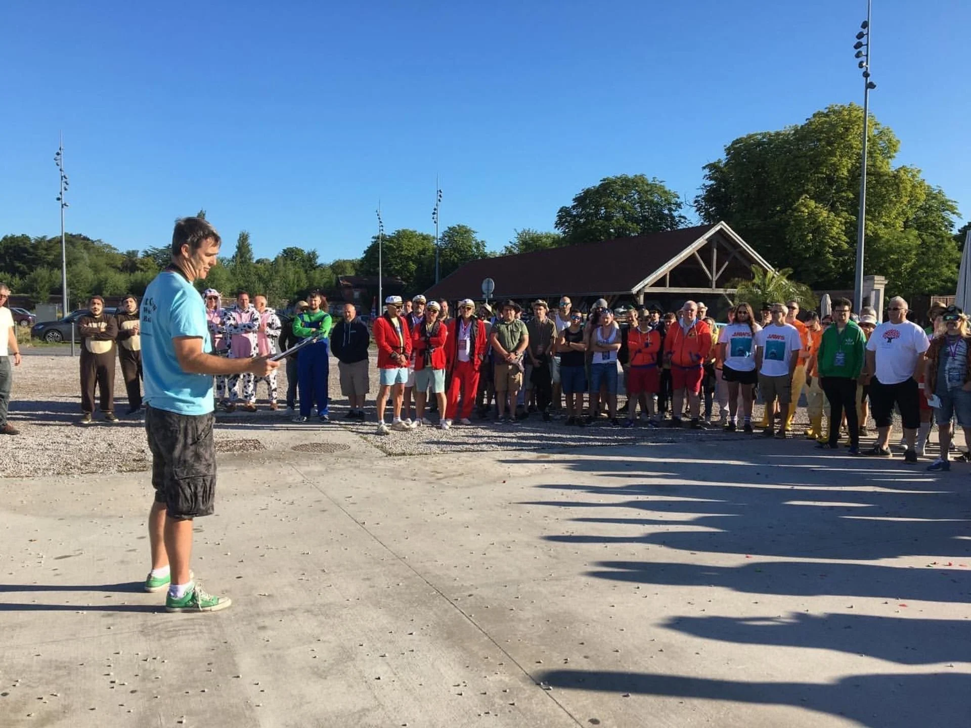A young man standing on a concrete surface reading a paper address a large group of people outdoors on a sunny day, with a small building and lush green trees in the background.