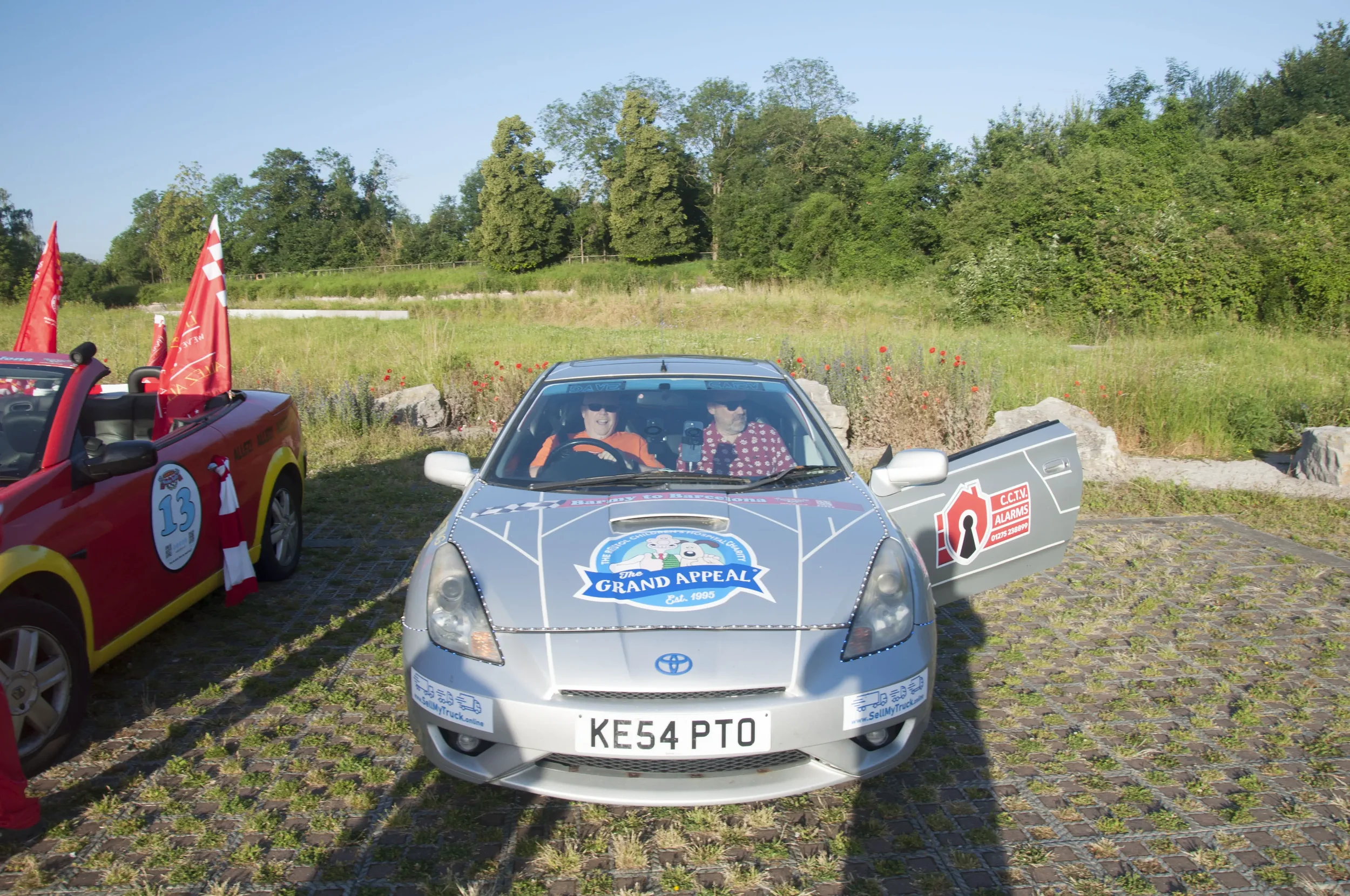 A silver Toyota car decorated with a 'Grand Appeal' logo on the hood, parked outdoors on a cobblestone surface, with two people inside. The car door on the right is open, and two women are sitting inside, one in an orange shirt and the other in a red