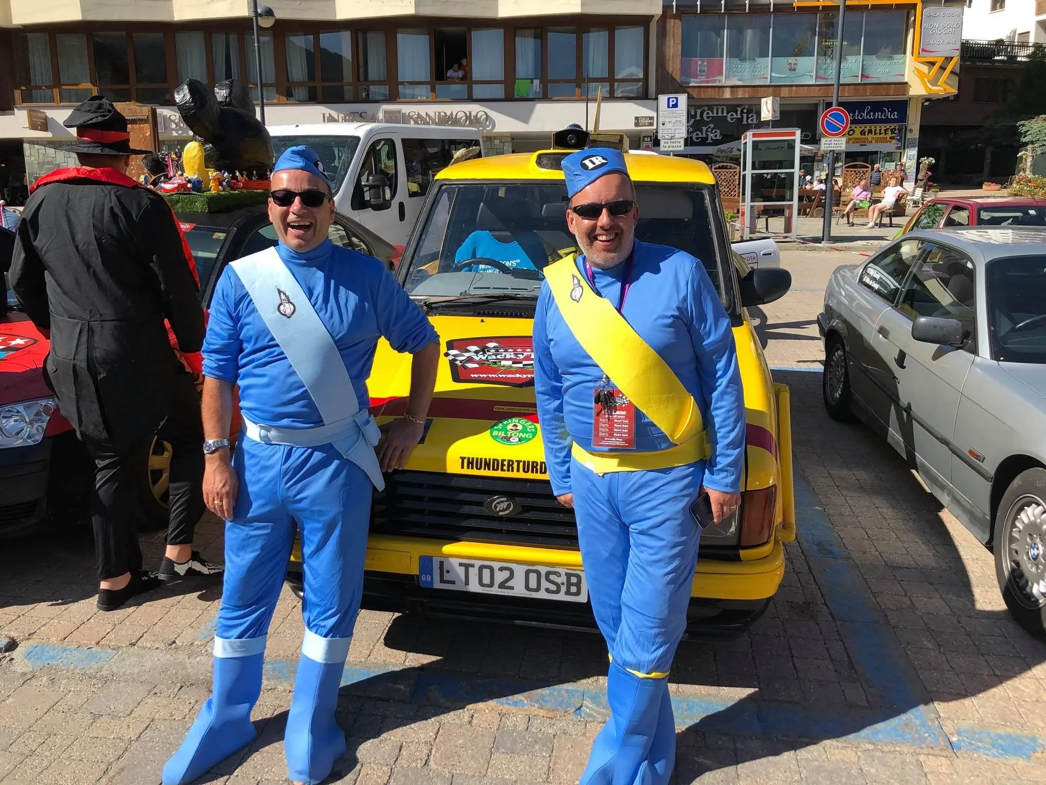 Two men dressed in blue racing costumes with yellow and light blue sashes, wearing blue hats and sunglasses, standing in front of a yellow car at an outdoor event. They are smiling and posing for the photo.